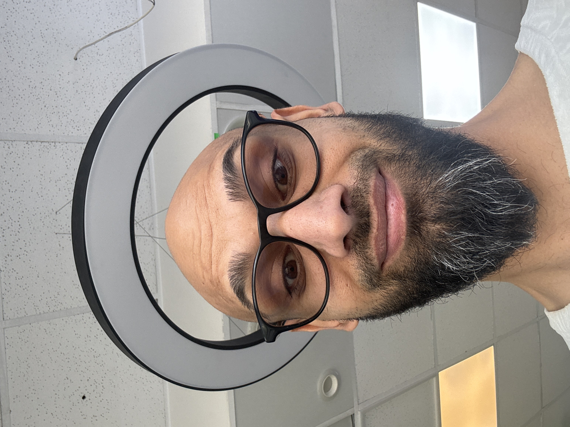 A close-up, slightly upward-angled portrait of a light-skinned man with a shaved head and a dark beard with some grey hairs, wearing black-rimmed glasses. He is looking directly at the camera. A ring light surrounds his head. The background is a white tiled ceiling with fluorescent lights. There is no indication of the location being in Plovdiv, Bulgaria.
