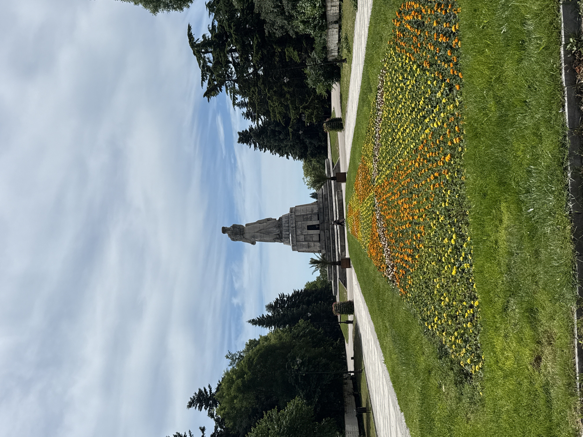 A wide-angle, eye-level outdoor shot captures a monumental stone statue on a tiered base, set against a backdrop of a partly cloudy sky and lush green trees. In the foreground, a meticulously maintained flower bed showcases rows of vibrant yellow and orange blossoms. A paved walkway borders the flower bed, leading towards the monument. The scene is bathed in natural daylight.