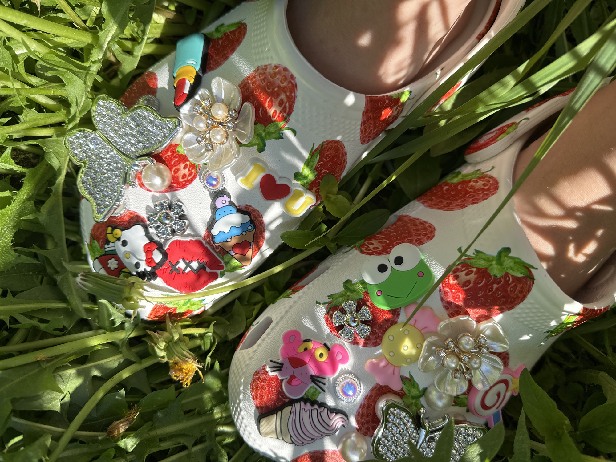 The image shows two white shoes decorated with strawberry and various cartoon-themed charms, resting in a bed of green grass and weeds. The shoes are adorned with charms like strawberries, butterflies, flowers, a frog, a pink panther, a cupcake, lips, a manicure stick, and an "I heart U" message. The location of the photo is not provided, so I cannot confirm if it is in Nizhny Novgorod, Russia.