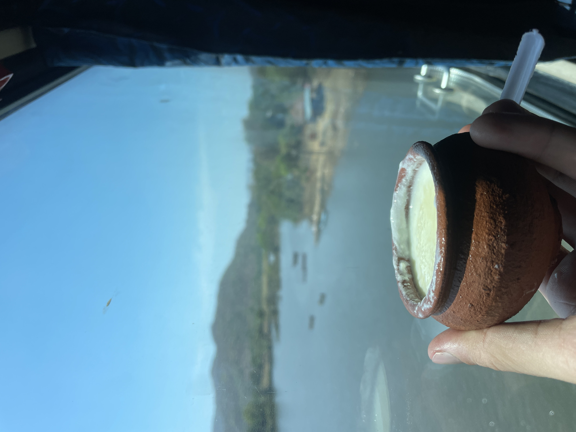 The image depicts a person holding a small, brown clay pot filled with white yogurt and a white plastic spoon. The yogurt appears thick and creamy. The background is a blurred landscape, possibly a view from a vehicle window, with a large body of water and a hill or cliff covered in greenery. The reflection in the window suggests the location may be in Dasgaon, India, based on similar landscapes in that area. There are boats on the water.