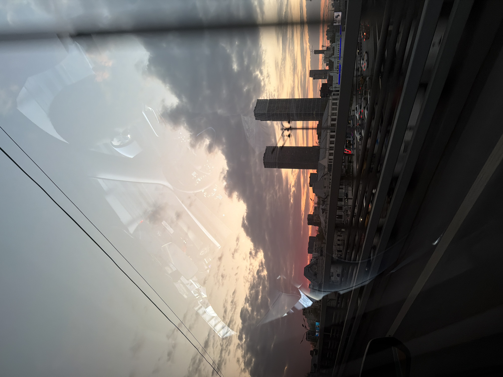 The image, taken from inside a car, shows a sunset view of a city skyline reflecting on the windshield. The reflection obscures the view, but it is possible to discern two tall buildings in the foreground, likely located in Moscow, Russia. The sky is filled with dark clouds and a vibrant sunset featuring orange and pink hues. The car's interior is visible as a reflection on the windshield. The edge of the bridge is seen on the right. The location is the city of Moscow, Russia.