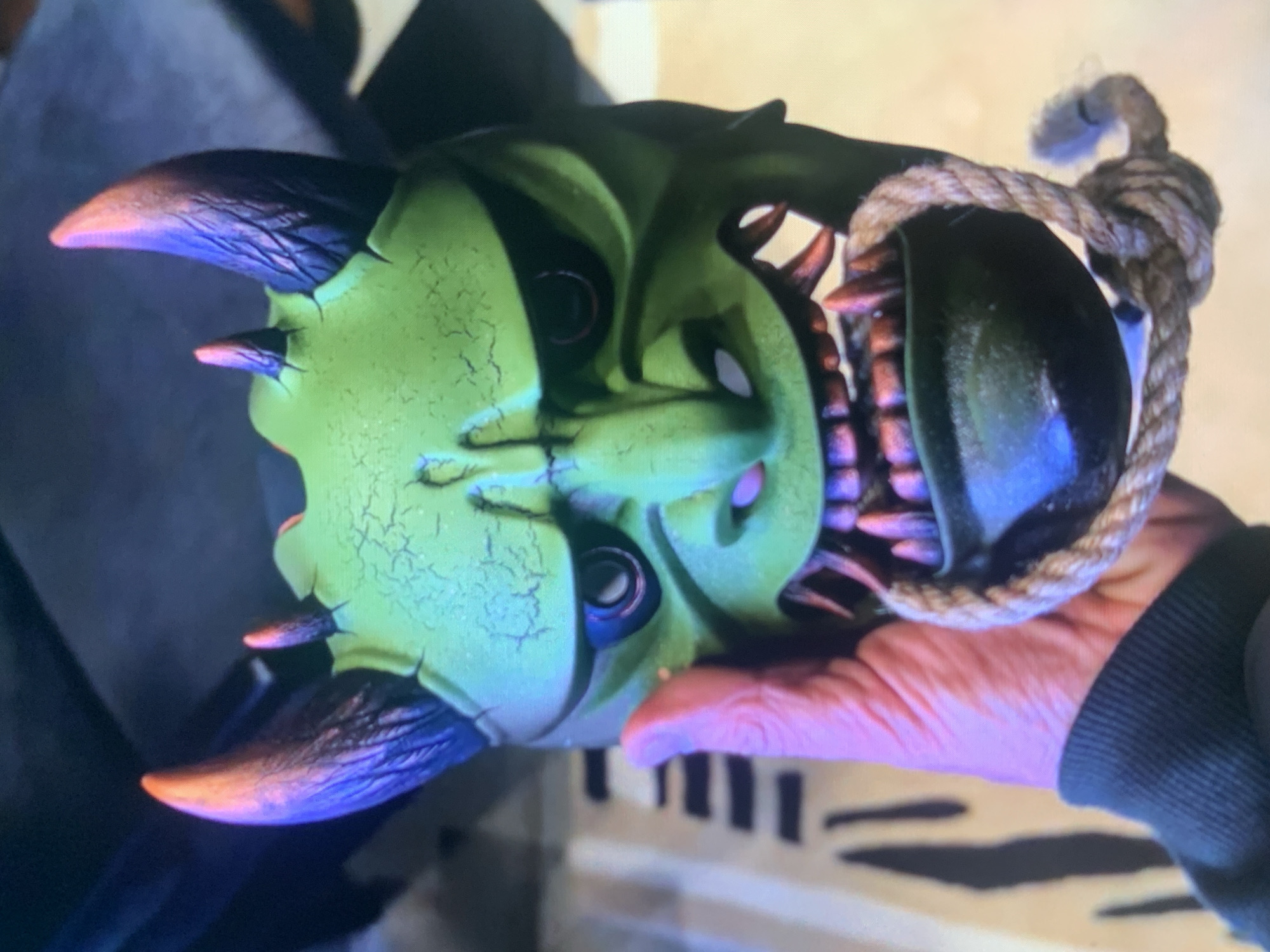 A hand holding a green, demonic mask with large horns and sharp teeth. The mask has a rope tied around its lower jaw. The background is blurred, but appears to be indoors with some dark objects.