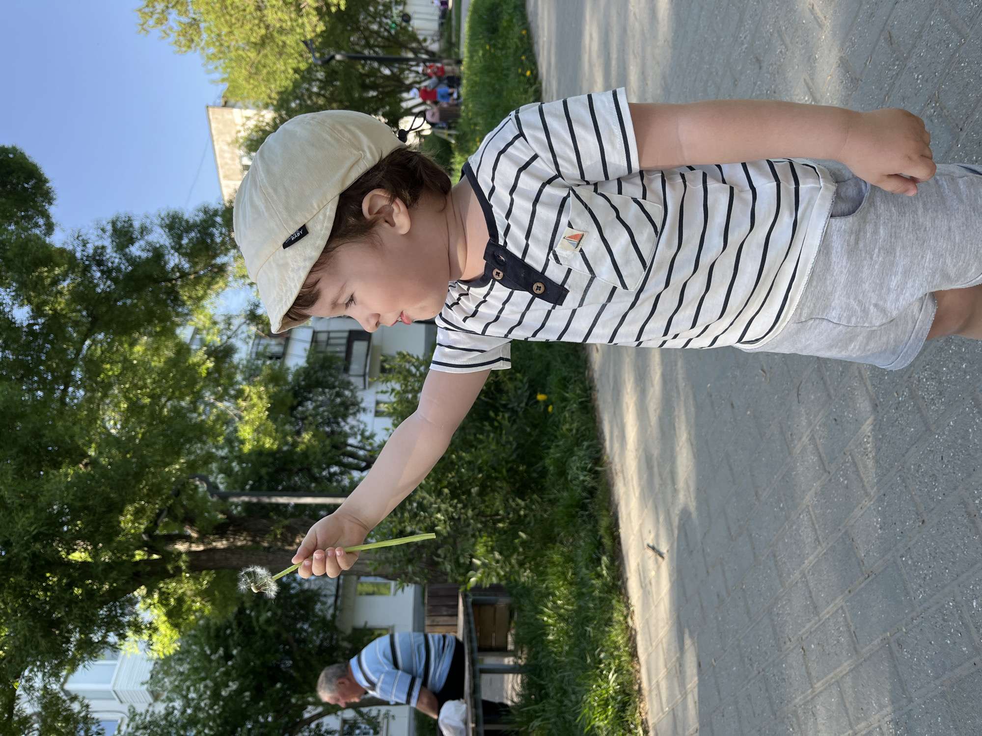 A young child wearing a beige cap, a striped shirt with a small sailboat insignia, and gray shorts holds a dandelion in their outstretched hand. They are standing on a paved sidewalk. A man in a blue and white striped shirt is bent over near a bench behind a patch of grass. Green trees and a building are visible in the background. The photograph appears to have been taken outdoors in a public space. While there is insufficient information to confirm the location, if the city is Chaikovsky, Russia, it would be relevant that it is a municipality and the location is likely a park or public green space.