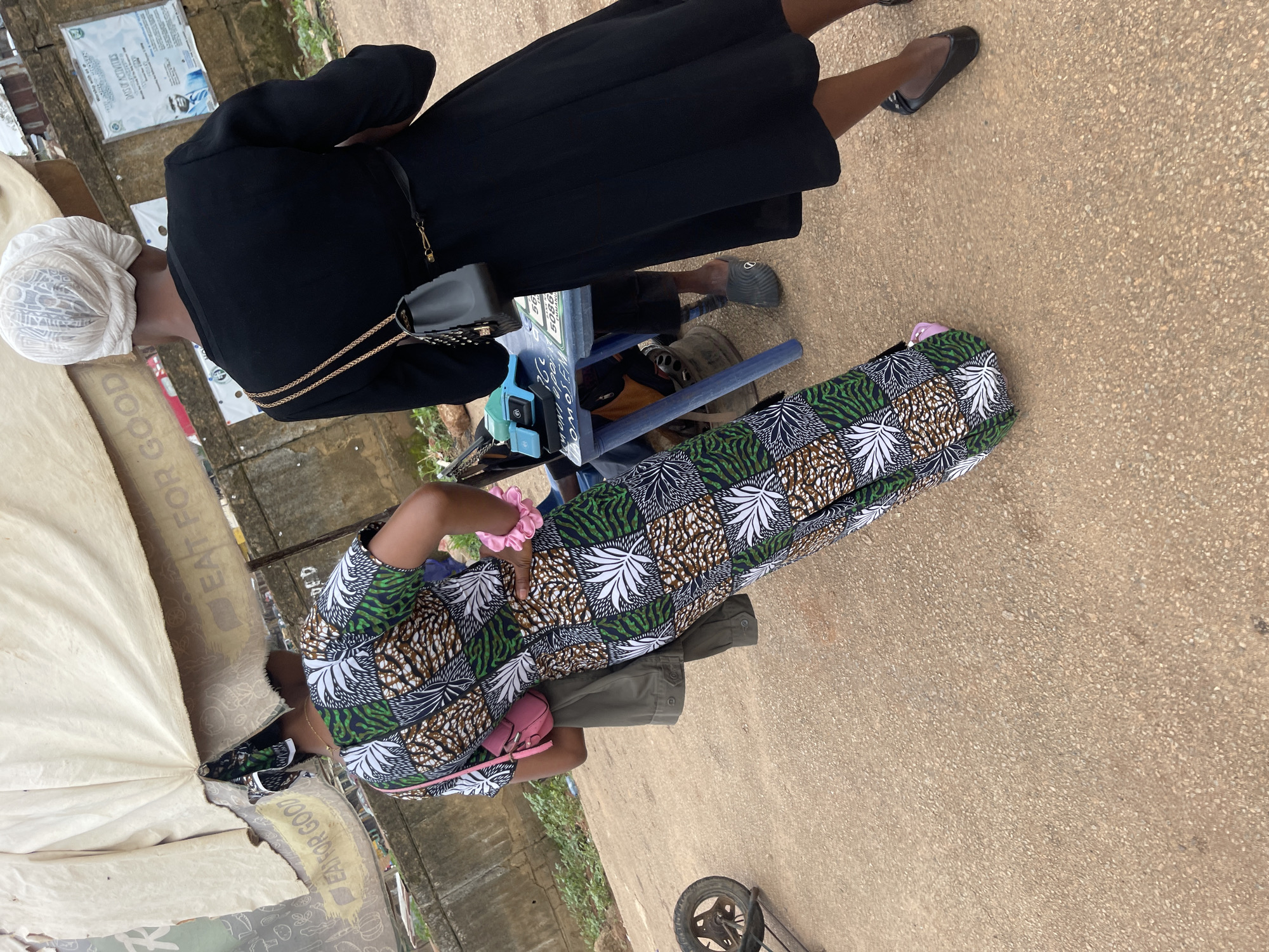 This image captures a street scene, likely in Rafin Par, Nigeria, where two women are engaged in what appears to be a transaction at a roadside stall. The woman on the left is wearing a vibrantly patterned long dress and a headscarf, and is seen from the back, her left hand resting on her hip. A pink purse is slung across her shoulder. The woman on the right is dressed in a black abaya-style dress and a white head covering, also viewed from behind. She has a small black handbag with a gold chain strap.
In front of the woman in black, a blue table holds a small, light-blue electronic device, possibly a scale, and some papers. The setting appears to be an outdoor market or street-side vendor area, indicated by the worn pavement, a dirt wall in the background adorned with posters, and a weathered tarp or awning overhead, which has text on it, partially obscured, including the phrase "EAT FOR GOOD." A rusty wheel attached to a metal frame is visible on the left side of the frame, suggesting the presence of carts or other mobile market equipment. The overall atmosphere is that of a bustling, everyday scene in a Nigerian town.