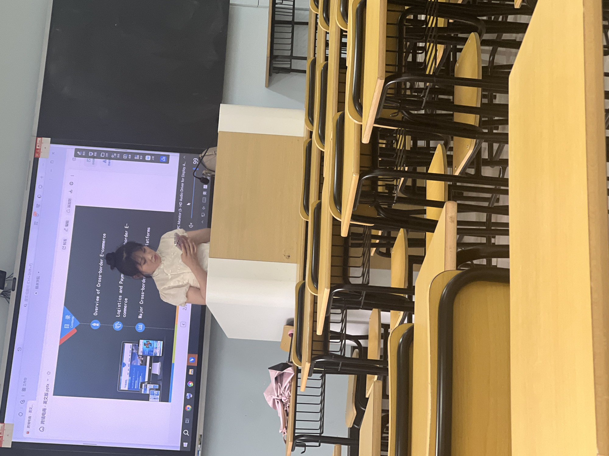 The image captures an indoor classroom setting with a large interactive display screen at the front. A young woman, dressed in a light-colored, high-necked top, is standing behind a podium in front of the screen, looking down at a smartphone she is holding in her hands. The screen displays a presentation slide with bullet points and graphics related to "Cross-border E-commerce." Rows of empty yellow desks with black metal frames are visible in the foreground, suggesting the class is either about to begin, has ended, or is in session with students off-camera. A pink umbrella is neatly folded and placed on one of the desks to the left. The room appears to be a typical educational environment, possibly a lecture hall or classroom within an institution in Liandu District, China, given the presence of the Chinese interface elements on the display screen. The lighting suggests it is daytime.