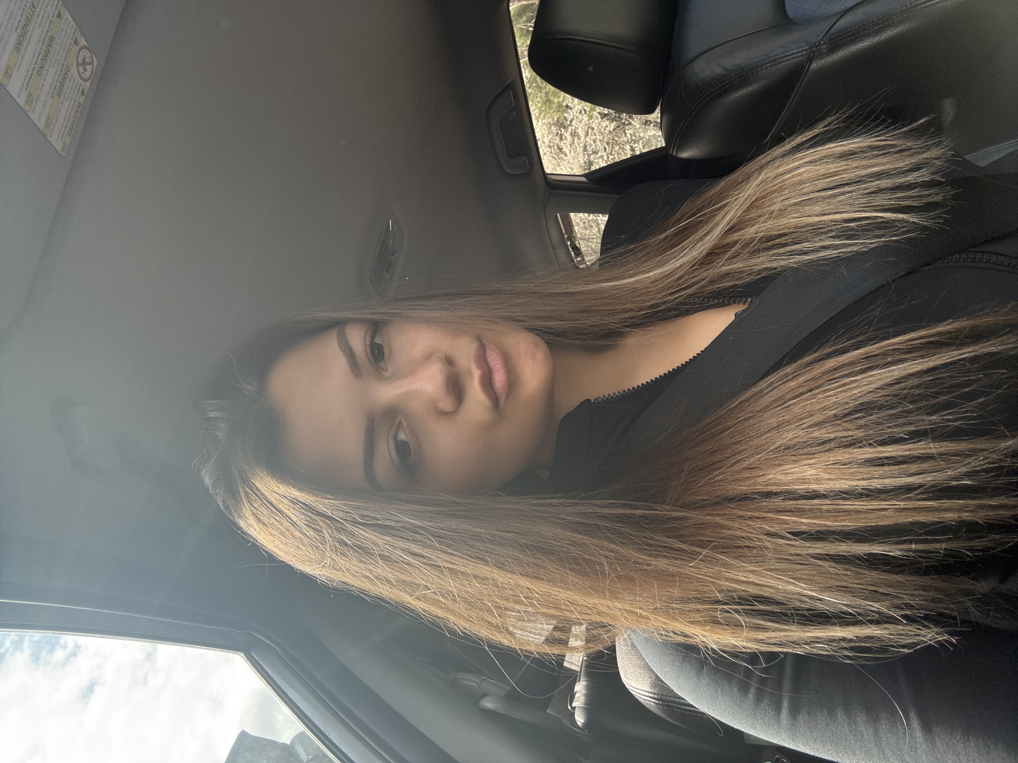 A woman is in the driver's seat of a car, wearing a seatbelt. She has long brown hair. The interior of the car is dark. The background visible through the car windows is out of focus, but appears to be an outdoor scene with some trees. The location is unknown, and there is no evidence indicating the city of Sofia, Bulgaria.