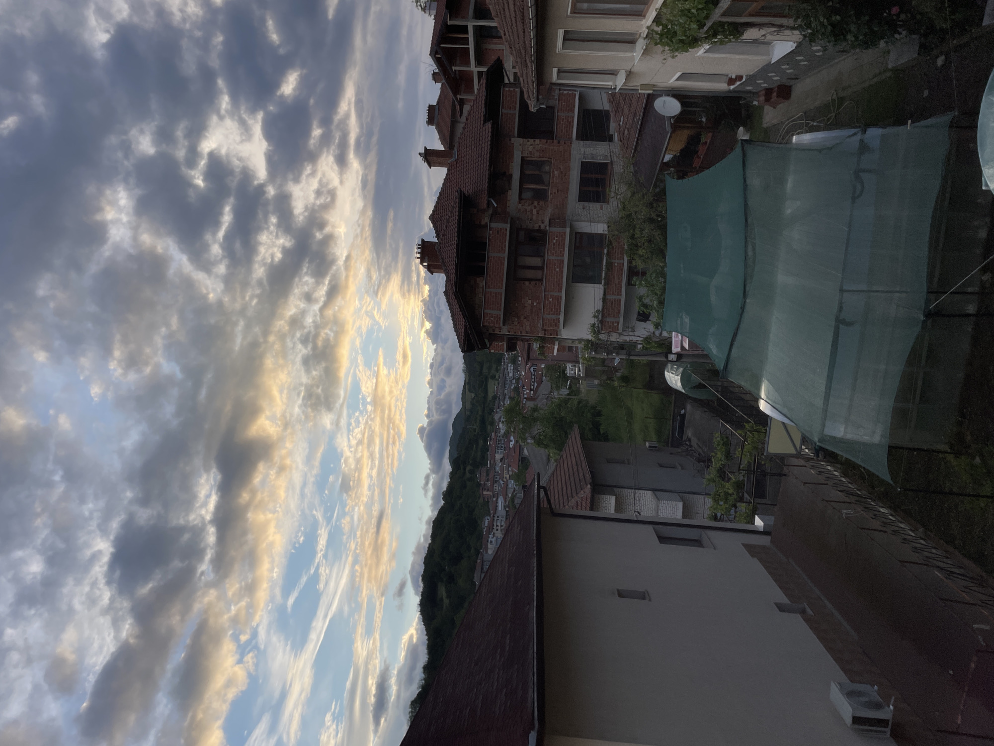 The image shows a view in Zlatograd, Bulgaria, featuring a cloudy sky with some patches of blue and hints of sunset colors. Below the sky is a residential area with various buildings of different heights and designs. One building has exposed red brick walls. There is green vegetation visible, including trees and possibly gardens. A green mesh or tarp-like structure is also visible. In the distance, there are mountains and more of the town nestled in the landscape.