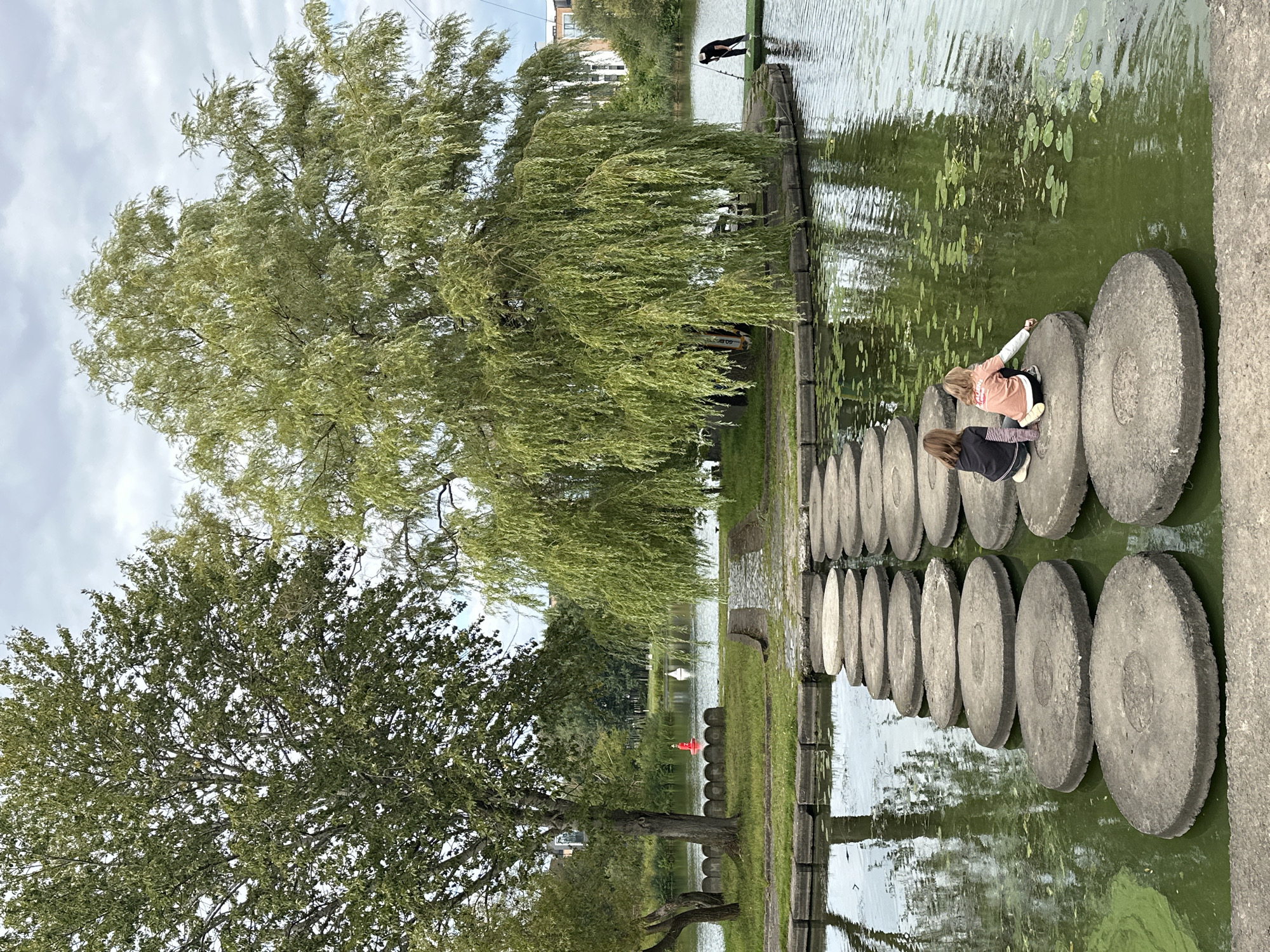 In Minsk, Belarus, two young girls are seen sitting on large, circular stepping stones placed in a tranquil body of water. The girl on the left, wearing a dark top with striped sleeves, is facing the water. The girl on the right, in a light orange top, is reaching towards the water with her hand. They are part of a pathway of these stepping stones that crosses the water. In the background, a large weeping willow tree with its branches cascading down provides shade, and a dense green tree stands to its left. The water is calm, reflecting the trees and the overcast sky. Further in the distance, a man in dark clothing is visible on a platform near the edge of the water, and a red and white buoy are floating in the lake. Buildings can be seen in the far background, suggesting an urban setting.