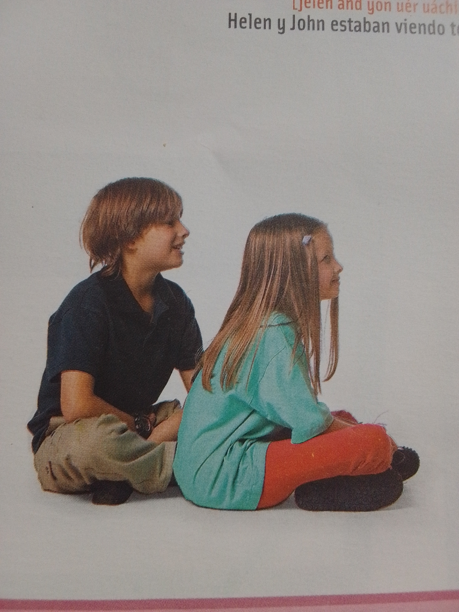 Two children, a boy and a girl, are sitting cross-legged on a plain, light-colored surface that could be a floor or a studio backdrop. The boy, on the left, has short brown hair and is wearing a dark blue collared shirt and khaki pants. He is looking to his right with a slight smile. The girl, on the right, has long blonde hair with a light blue clip and is wearing a turquoise shirt and orange pants. She is looking to her right, also with a slight smile. The background is a solid, light gray color with some faint shadows. There is text at the top of the image that reads "Helen y John estaban viendo te". The scene appears to be staged, likely for a photograph or illustration. There is no indication or visual information suggesting the location is in La Tagua, Venezuela.
