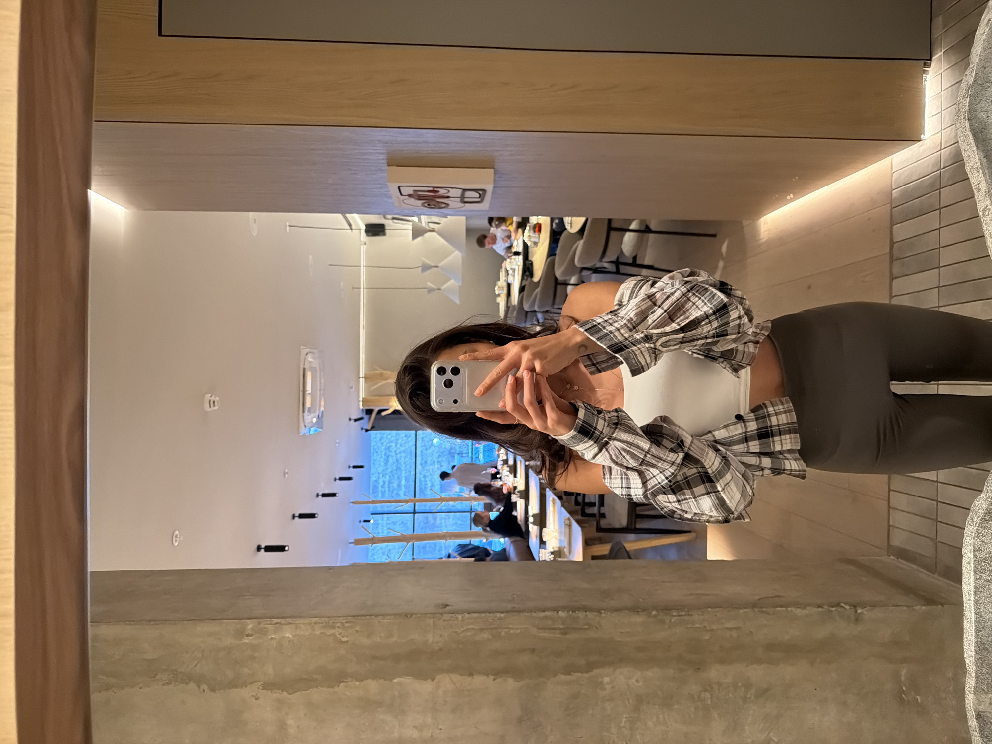 A person is captured in a mirror selfie within a modern interior, likely a restaurant or hotel lounge. The central subject, appearing to be a woman, holds a silver iPhone (possibly a Pro model) with multiple cameras in both hands to take the picture. She is dressed in a white crop top, an open brown, black, and white plaid shirt, and dark grey leggings. Her long, dark hair frames her face, and a subtle tattoo is visible on her wrist.
The reflection in the mirror reveals a spacious and contemporary dining area behind her. Several patrons are visible seated at tables, with one individual in a white shirt particularly clear in the background. The interior design features light-colored wooden panels on the ceiling and walls, complemented by integrated strip lighting that casts a warm glow. Modern tables and chairs furnish the space. A small, white rectangular piece of artwork, displaying abstract red and black circular designs, is mounted on one wooden panel. A large window, reflecting a bright blue hue suggestive of the sky or an outdoor feature, is prominent, near which minimalist black stick-like decorative elements or light fixtures are arranged on a wall.
The mirror is bordered by a vertical wooden panel on the left and a horizontal wooden beam at the top. The bottom portion of the image shows a textured, light grey concrete-like surface, likely the floor or a base structure. To the far right, a section of a grey tiled wall with a vertical light strip is visible, alongside a large, irregular grey stone or sculptural element. No explicit text is clearly legible within the image. The setting suggests an upscale and contemporary venue, consistent with a modern establishment in Krasnoyarsk, Russia.