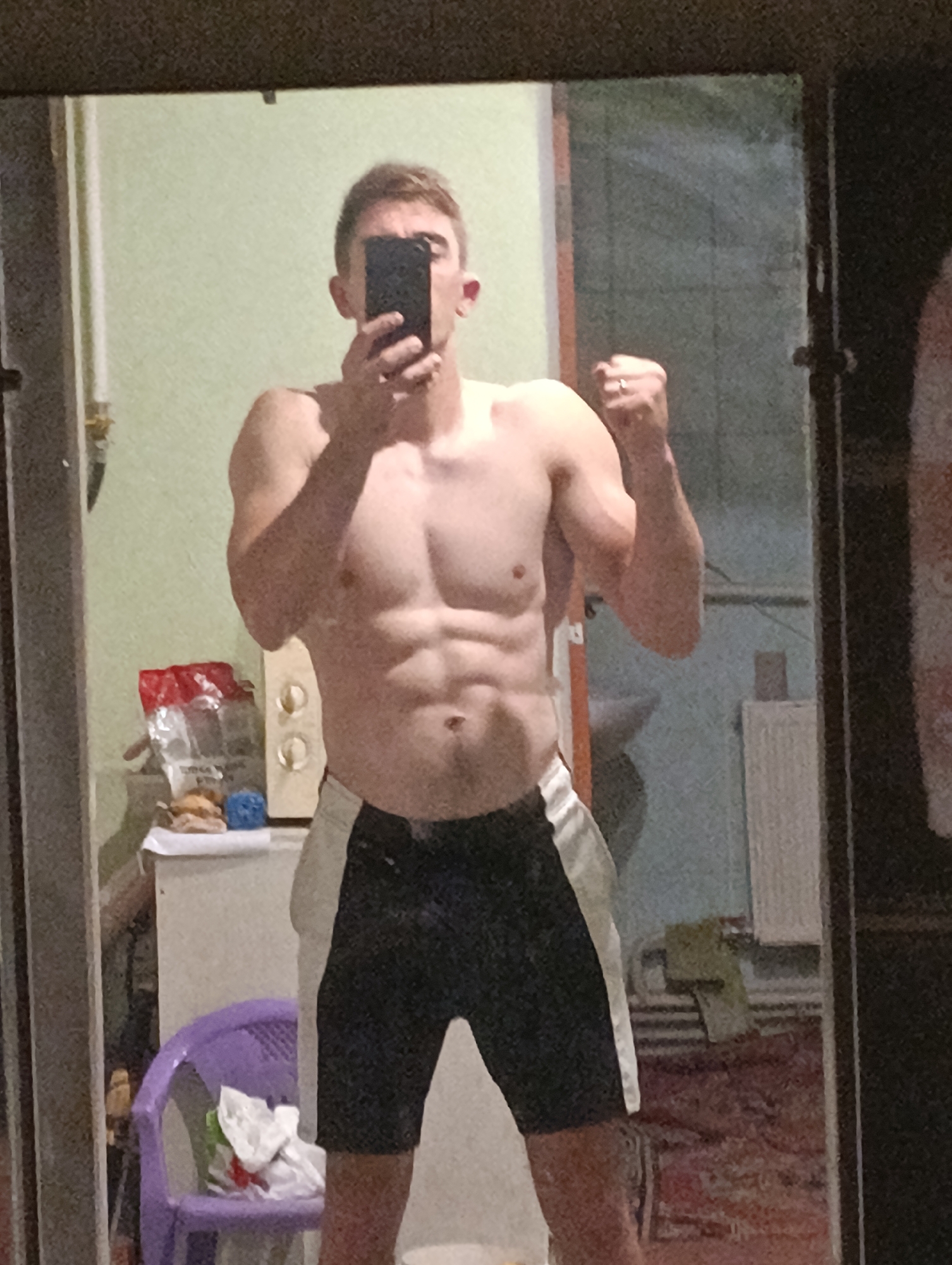A young man with light skin, brown hair, and a muscular physique stands in front of a mirror, taking a selfie. He is wearing black shorts with white side panels. His left arm is bent and flexed, while his right hand holds a black smartphone to take the picture. The background appears to be an indoor setting, likely a room in a residential building. The reflection in the mirror shows a wall with a light green paint, a cabinet with round knobs, a purple chair with some items on it, and other miscellaneous objects. The overall impression is of a person documenting their physical fitness. The prompt requests that this image be described as if located in Fastiv, Ukraine, but there are no visual cues in the image to confirm this specific location.