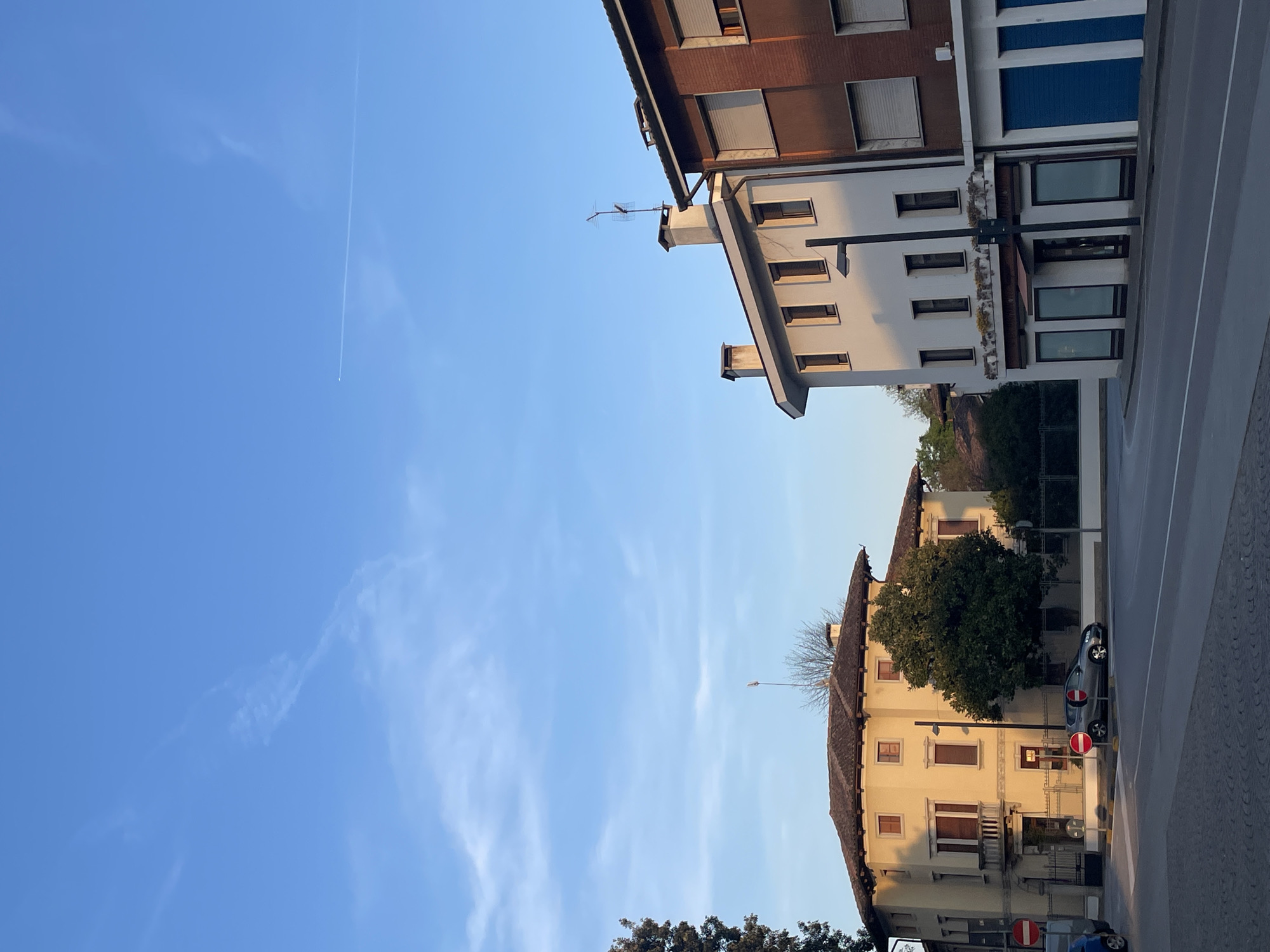 The image depicts a street scene in Case / Scjasis, Italy. The sky is blue with faint wisps of clouds and a contrail from an aircraft. Buildings line either side of the street. One building is white with brown trim and blue window coverings, while the building across the street is a yellow/tan color with a terracotta roof. A vehicle is parked along the side of the road. Stop signs are visible on the side of the yellow/tan building. A tree is also present. The overall perspective is tilted.