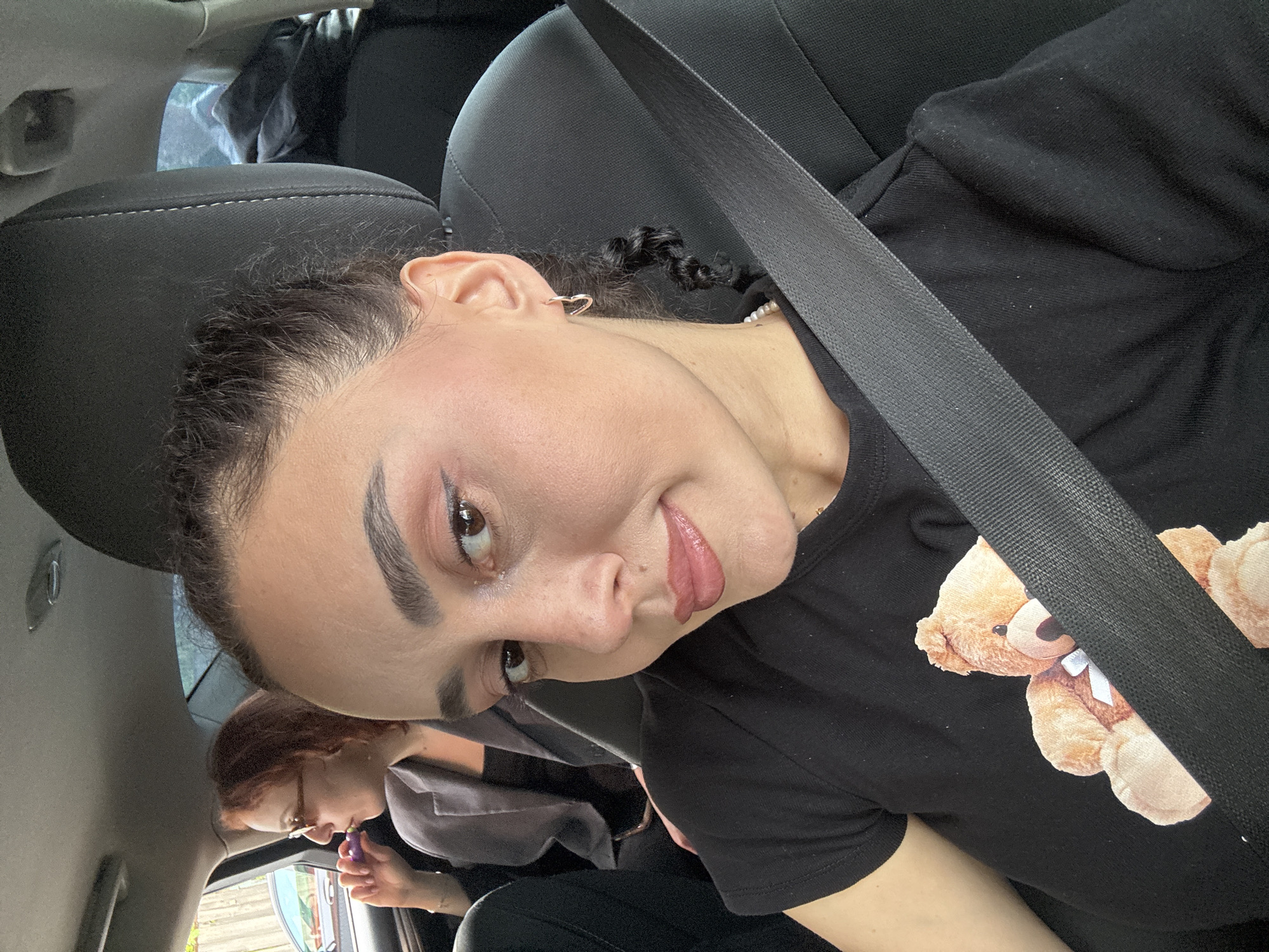 A woman wearing a black shirt with a teddy bear graphic is seated in a car, buckled up. Another person is visible in the backseat. The location is unknown.