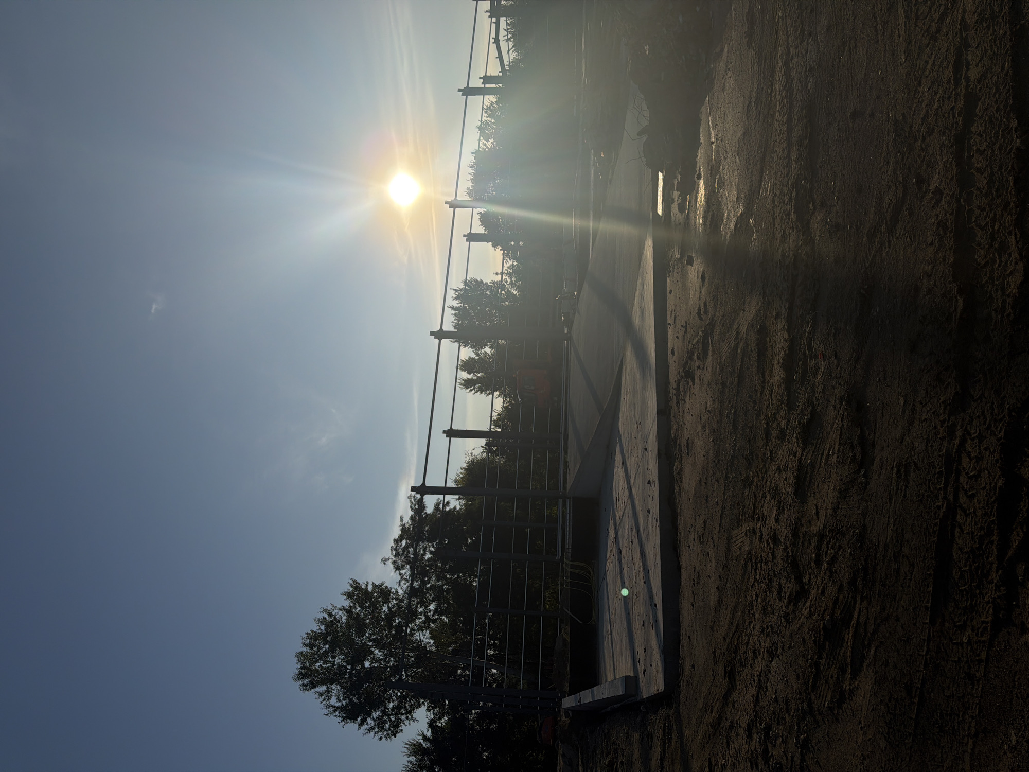 The image depicts a construction site bathed in the warm glow of the setting sun, located in Vatra, Moldova. A concrete ramp leads up to a metal railing structure, possibly part of a bridge or walkway under construction. The ground in the foreground is unpaved and disturbed, showing tire tracks from machinery. Trees line the background, silhouetted against the bright sky. The sun is low on the horizon, casting long shadows and creating lens flare across the scene. An orange construction vehicle is partially visible on the left side of the ramp. No people are visible in the frame.