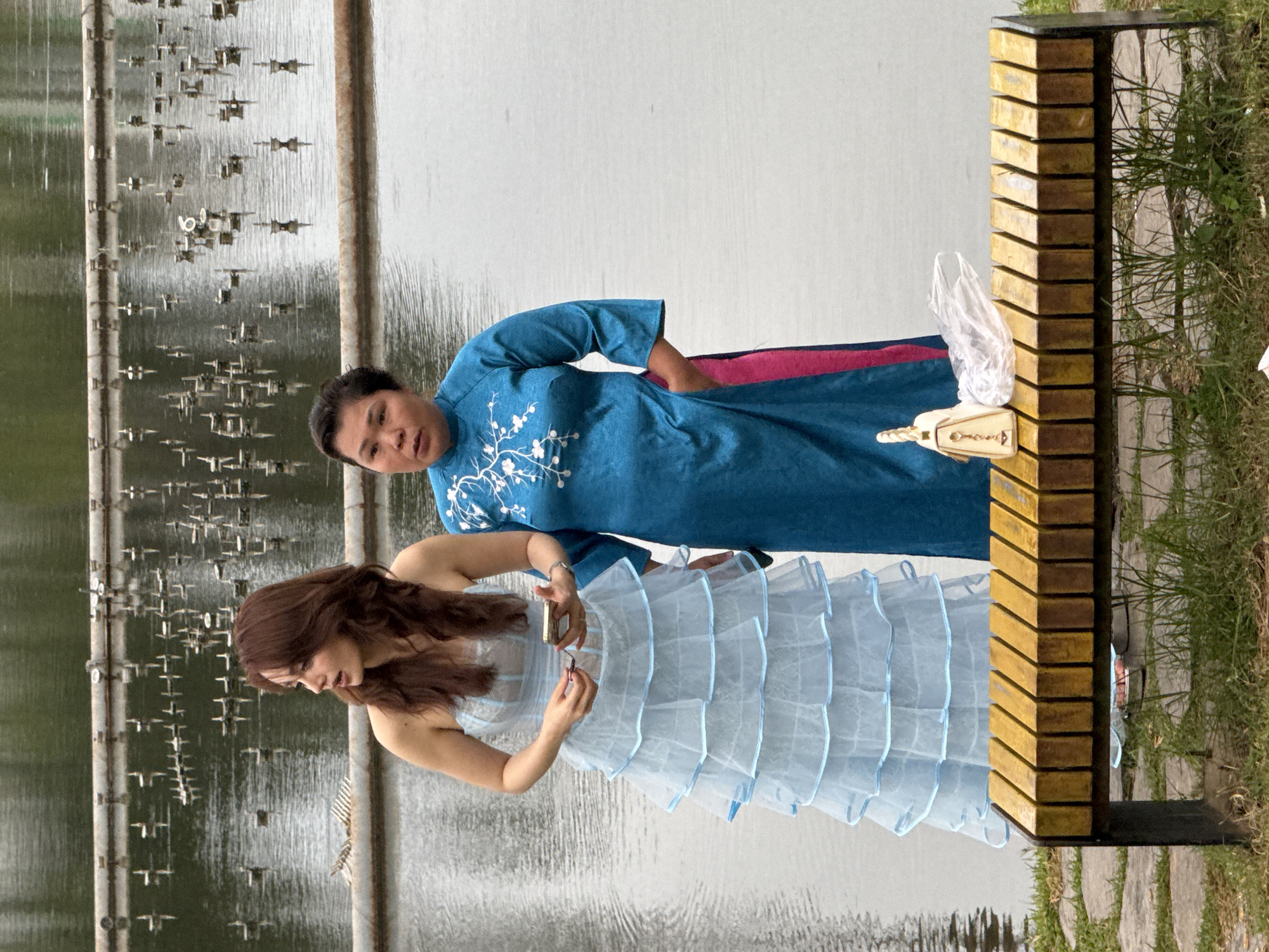 In the city of Da Lat, Vietnam, two women stand near a water feature. The woman on the left, with long, dark brown hair, wears a strapless, tiered light blue dress adorned with ruffles and blue trim. She holds a smartphone in her right hand. To her right stands another woman with dark hair pulled back, wearing a vibrant turquoise dress with embroidered white floral patterns and pink trim. Her right hand is in her pocket. In front of them, a wooden slatted bench holds a cream-colored purse and a crumpled plastic bag. The background features a body of water with numerous small, metallic structures emerging from it, likely part of a fountain system. The area around the bench is covered in grass and stone paving.