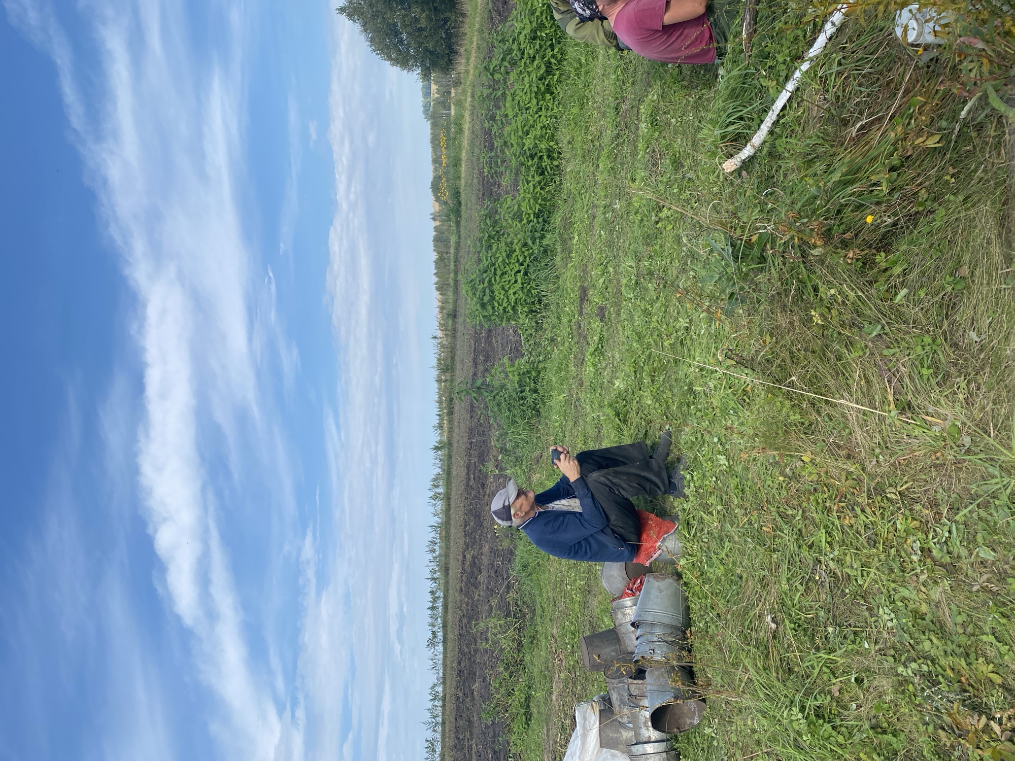 In Tigritskoe, Russia, a man wearing a blue jacket, black pants, and a gray cap sits on a red cloth covering a bucket, looking at his phone. He is surrounded by several metal buckets and agricultural fields. The sky is blue with white clouds. Another person, partially visible and wearing a pink shirt, is also present in the scene.