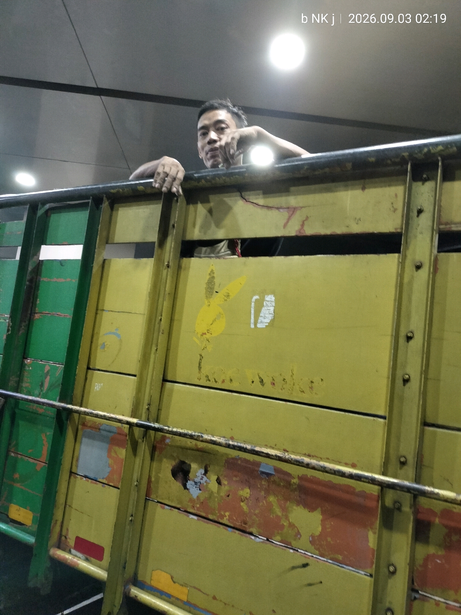 A man, appearing to be of Southeast Asian descent, is visible peering over the edge of a yellow truck bed. He is wearing a dark shirt and his hands rest on the metal railing of the truck. The truck bed is constructed from horizontal wooden planks painted yellow, with some sections showing wear and tear, revealing rust and peeling paint. A green section is visible on the left side of the truck. A yellow sticker with the Playboy bunny logo is affixed to one of the yellow planks. In the background, a ceiling with exposed beams and a bright light fixture are visible. Text overlay on the image reads "b NK j | 2026.09.03 02:19". The scene appears to be outdoors at night, possibly in a parking lot or loading dock area, suggesting a location in South Jakarta, Indonesia, given the context provided.