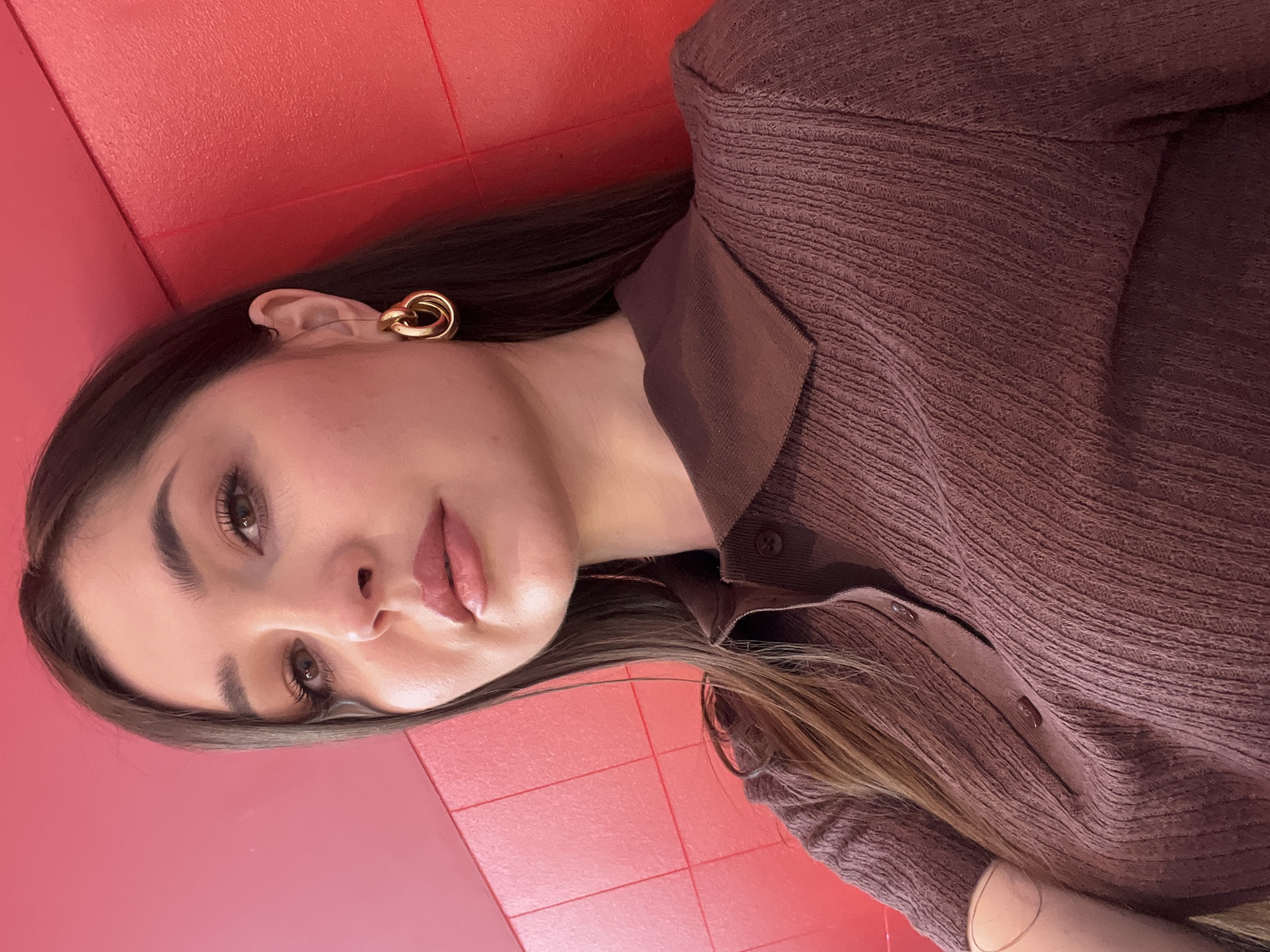 A close-up portrait features a young woman looking directly at the viewer, set against a vibrant red background. The image is originally oriented vertically, with her head to the left and body extending to the right.
The woman has long, straight, dark brown hair, which falls past her shoulders. She has fair skin, well-defined dark eyebrows, and brown eyes accentuated by mascara or eyeliner. Her lips are full and tinted with a light pink or nude lipstick, slightly parted. On her visible right ear, she wears a large, gold-toned earring shaped like intertwined loops.
She is dressed in a brown, long-sleeved top or sweater. The fabric has a distinct textured, ribbed pattern. The garment features a collar and a partial placket with visible buttons down the front.
The background is a solid, vivid red or coral color, which appears to be a wall, possibly tiled, given the subtle rectangular lines visible in some cropped sections. The lighting is even, suggesting an indoor setting. There are no other visible objects, text, or discernible location cues within the frame. The woman's expression is neutral to serene.