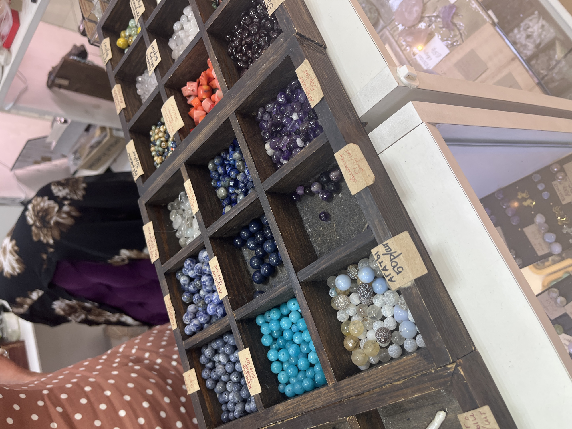 The image shows a close-up of a wooden tray filled with various types of beads, likely in a market or shop. The beads are sorted into separate compartments within the tray. Some compartments contain blue, purple, and teal colored beads, while others hold lighter, multi-colored beads and translucent white beads. Small paper labels with handwritten text, appearing to be prices and names of the beads in Russian, are attached to some compartments. In the background, parts of people's clothing and other displays are visible, suggesting a retail environment. The overall scene is well-lit, highlighting the textures and colors of the beads. The city of Sochi, Russia, is not explicitly depicted or mentioned in the image.