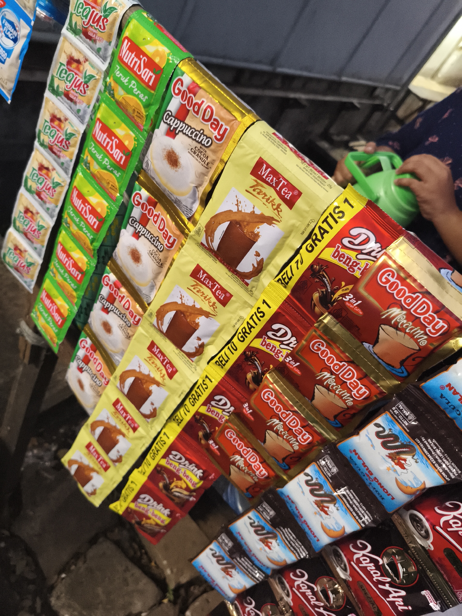 This image captures a vibrant display of various instant beverage sachets, likely at a street stall or small shop in Cibinong, Indonesia. The sachets are neatly arranged in vertical rows, hung for sale.
The visible products include instant tea ("Teajus" Gula Batu), instant orange drinks ("NutriSari Jeruk Peras"), instant coffee mixes like "Good Day Cappuccino" (with Cocoa Granule) and "Good Day Mocaccino" (3in1 Instant Coffee), and "MaxTea Tarikk" (an instant milk tea). Also present are "Dring beng-beng" chocolate malt drinks, "Liong Bulan Gula" (sugar), and "Kapal Api Special" coffee. A "Frisian Flag" instant milk sachet is partially visible.
Many of the sachets feature promotional offers, such as "BELI 10 GRATIS 1" (Buy 10 Get 1 Free) and "BELI 70 GRATIS 1" (which appears to be a typo, likely intending "BELI 10 GRATIS 1"). Packaging details like "NET WT/BERAT BERSIH 25g" and "HALAL" are also legible, along with a production origin for "Liong Bulan" in Bogor, Indonesia.
In the blurred background, a person's hands are visible, holding a bright green object, possibly a toy or a small watering can, suggesting activity in the vicinity of the stall. The lighting indicates either daytime or artificial indoor illumination. The ground appears to be a rough, dark surface like concrete or pavement.