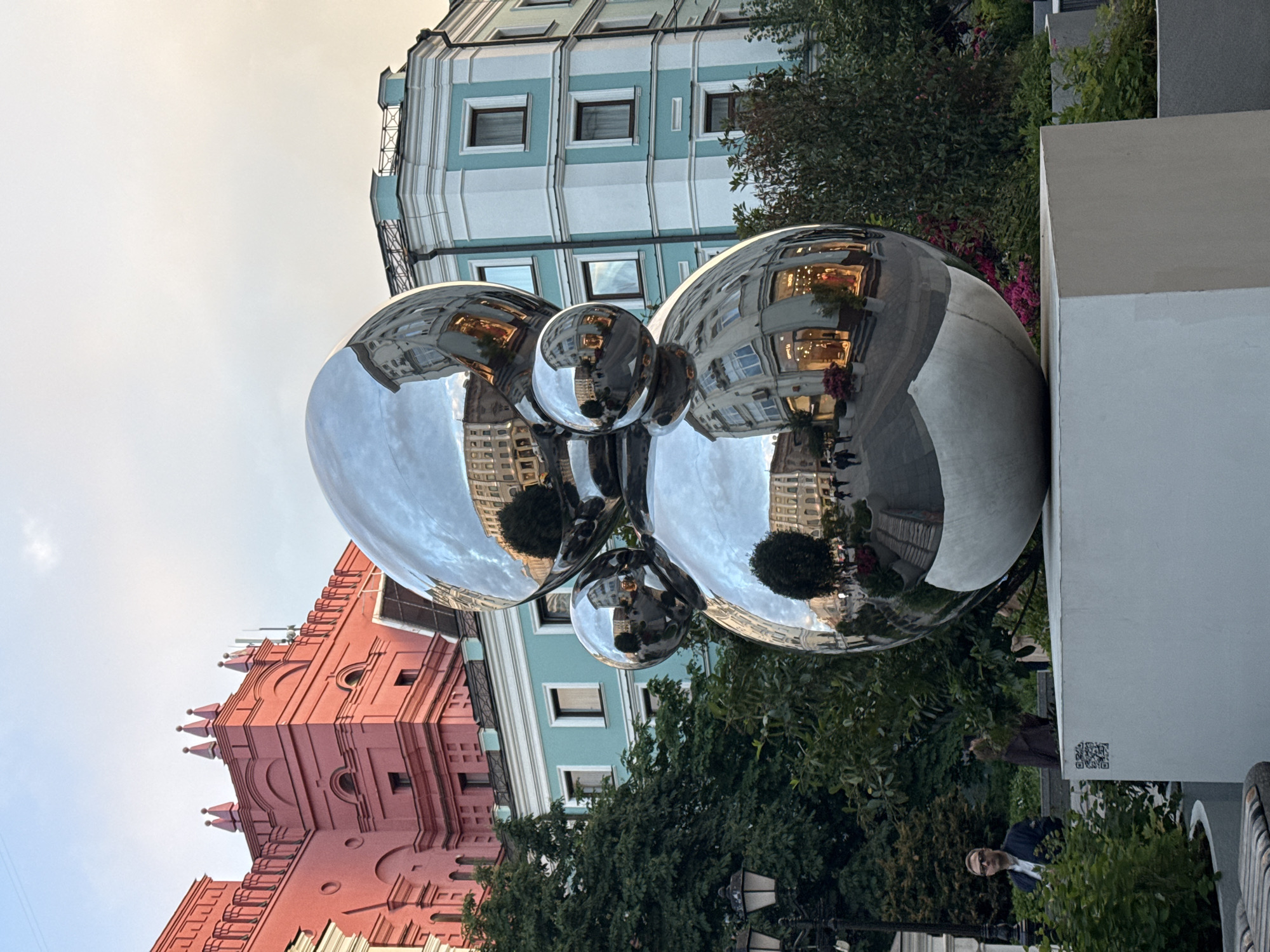 The image shows a reflective sculpture composed of interconnected spheres, positioned in an urban environment. The sculpture reflects the surrounding architecture. In the background, there is a red building and a building with a light blue and white facade, suggesting that this might be located in Moscow, Russia. The sculpture is situated on a gray and white pillar surrounded by greenery. At the bottom of the image, there is a person peering through the bushes to the right of the pillar.