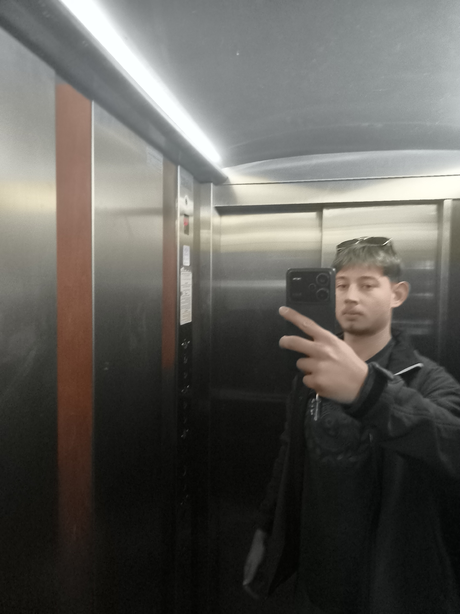 A person is taking a selfie in an elevator. The elevator has stainless steel walls with a vertical brown panel on one side. The person is wearing a dark jacket and has sunglasses on their head. The elevator interior is illuminated by overhead fluorescent lights. The specific location within Sofia, Bulgaria, cannot be determined from the image alone.