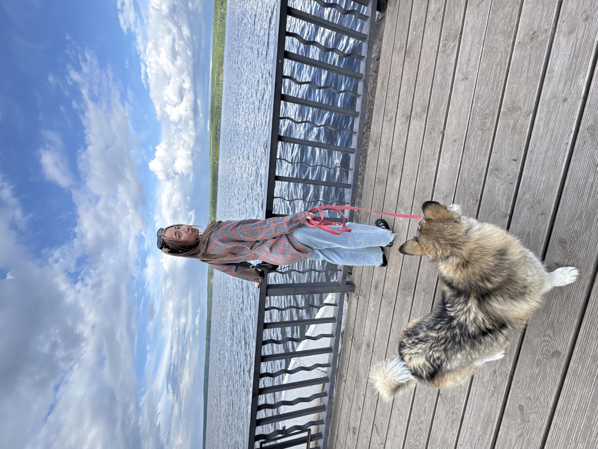 A young woman and a dog are on a wooden pier overlooking a body of water under a partly cloudy sky. The woman is leaning against a black metal railing, wearing a plaid jacket, light blue jeans, and dark sandals. She holds a red dog leash. The dog, with a multi-toned brown and black coat, stands on the pier near the woman. The body of water extends into the distance with a strip of green trees on the opposite shore. The pier is constructed of light-colored wooden planks. The location is in the city of Chaikovsky, Russia.
