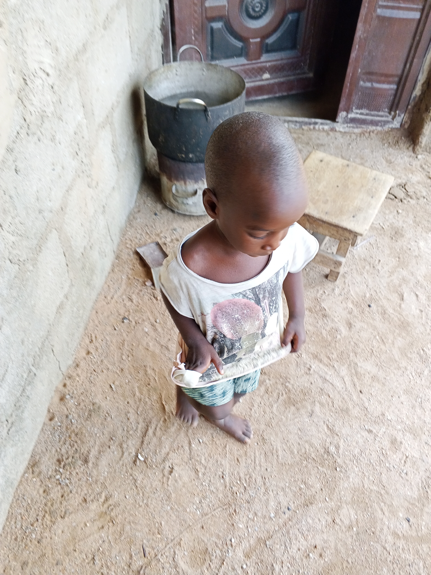 This image, taken in Adonkolo, Nigeria, captures a high-angle view of a young, dark-skinned child standing on sandy, earthen ground during daylight. The child, likely a toddler, has a closely shaved head and is barefoot. They are wearing a light-colored t-shirt with a complex graphic design featuring pinkish and dark tones on the front, and patterned shorts that are green and blue. The child is looking downwards, with their hands engaged in holding or examining the bottom hem of their t-shirt.
The setting appears to be an outdoor or semi-outdoor space adjacent to a building. To the left, a light-colored, textured wall, possibly made of concrete or mud brick, runs vertically. In the background, to the right, a dark brown wooden door with intricate carvings or panels is slightly ajar, revealing a dark interior. Next to the door and against the wall, a large, dark metal pot with two handles rests on a cylindrical metal base, suggesting a stove or cooking area. The pot appears to contain liquid. A small, light-colored wooden stool or bench is also visible in the background near the door. The ground is uneven, dusty, and strewn with small pebbles, typical of an unpaved area. The bright, natural lighting indicates it is daytime. No discernible text is visible on any objects.