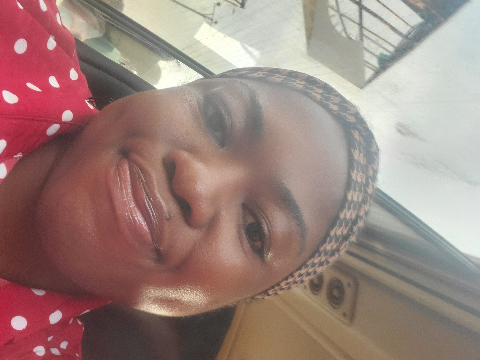 A young Black woman is taking a selfie inside a vehicle. She is wearing a red shirt with white polka dots and a patterned headscarf. Her head is slightly tilted, and she is smiling. The interior of the vehicle is visible, including a window with a reflection and the ceiling with a vent. The background suggests an urban environment, potentially Kakau, Nigeria, given the context.