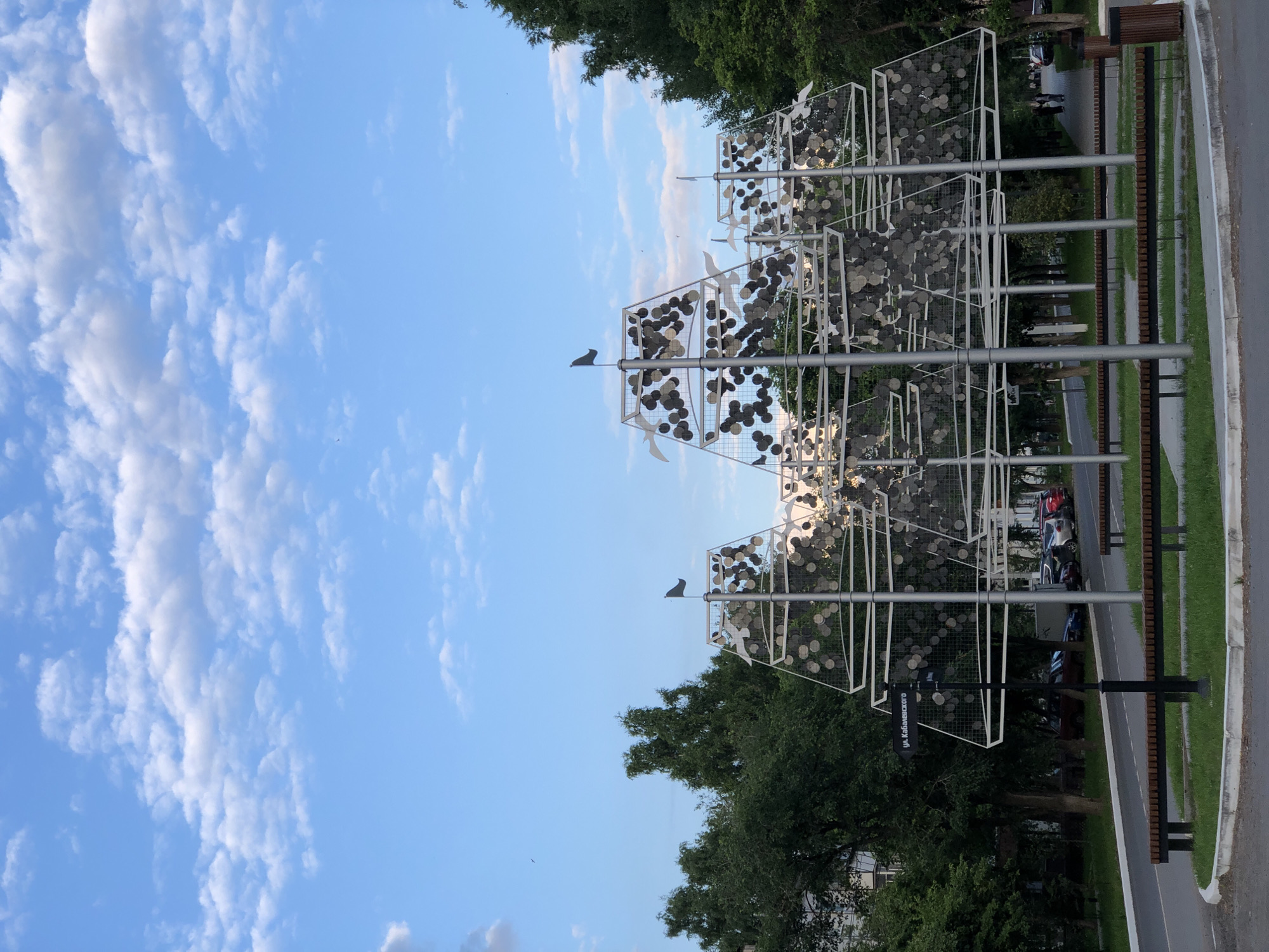 This media file captures an outdoor scene with a prominent art installation featuring white geometric structures adorned with numerous circular metallic discs. White bird-like figures are strategically placed on the installation, giving the impression of flight. The installation stands amidst lush green trees and a bright blue sky dotted with scattered white clouds, suggesting a pleasant daytime.
In the background, a paved walkway is visible, bordered by grassy areas and wooden benches, indicating a public park or promenade. A few cars are parked along the side of the walkway, and a street sign with Cyrillic text, "ул. Кабаненского", is mounted on a lamppost. The overall mood is serene and possibly reflects a calm, everyday moment in a public space. The lighting suggests it's either morning or late afternoon.