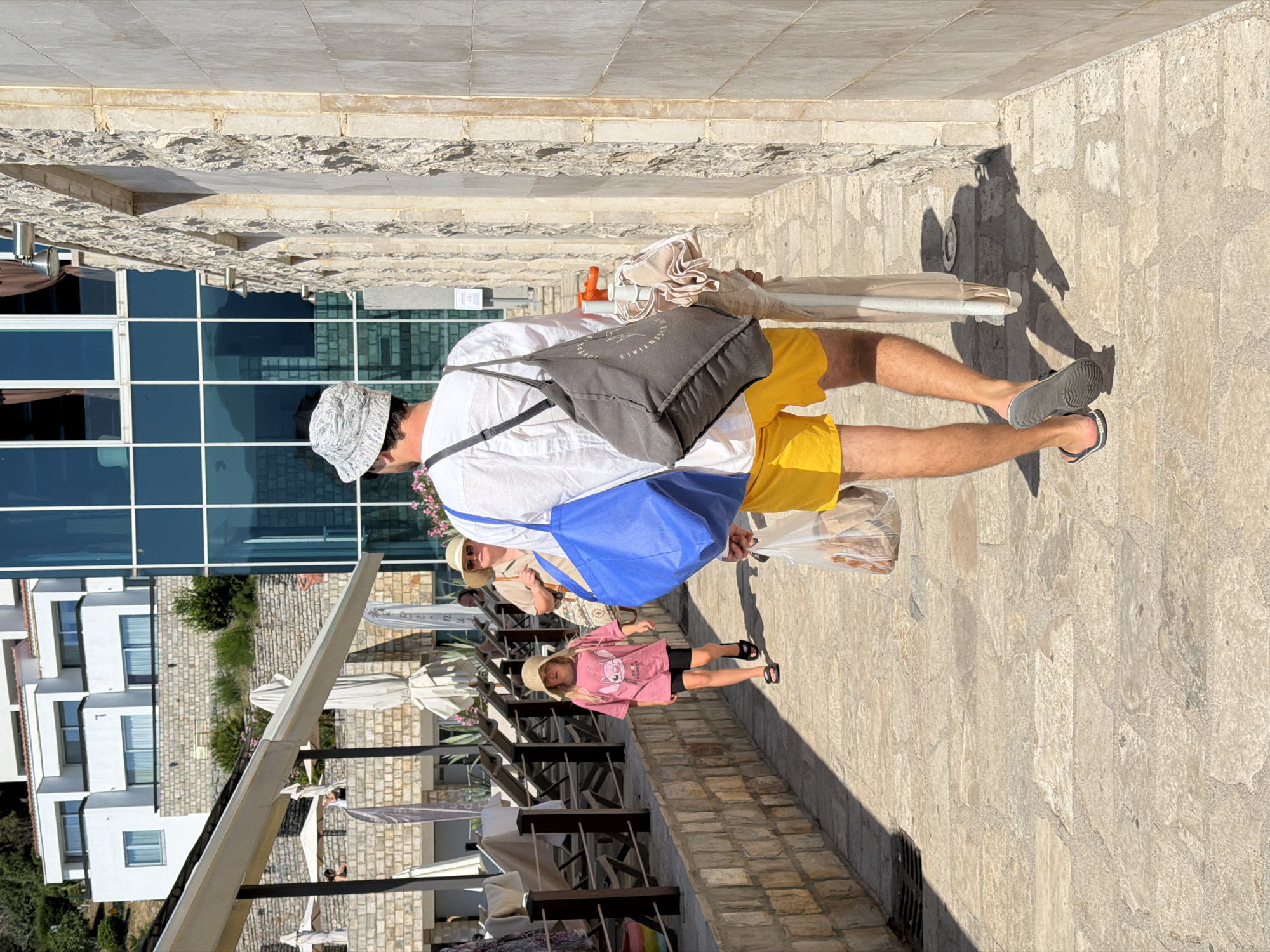 A man, a child, and another person are walking along a cobblestone path lined with stone buildings in Budva, Montenegro. The man in the foreground, wearing yellow shorts and a white shirt, carries a blue bag and a grey tote bag. He also holds a folded umbrella and a plastic bag. The child, a young girl in a pink t-shirt and black shorts, walks ahead of the man, wearing a straw hat and flip-flops. The third person, wearing a wide-brimmed hat and a light-colored top, is further back on the path, partially obscured. The environment includes a modern building with blue glass windows and a white building with multiple stories situated on a hillside. The area appears to be a tourist destination, with awnings and lounge chairs visible in the background. The lighting suggests it is daytime, with strong sunlight casting shadows on the path.