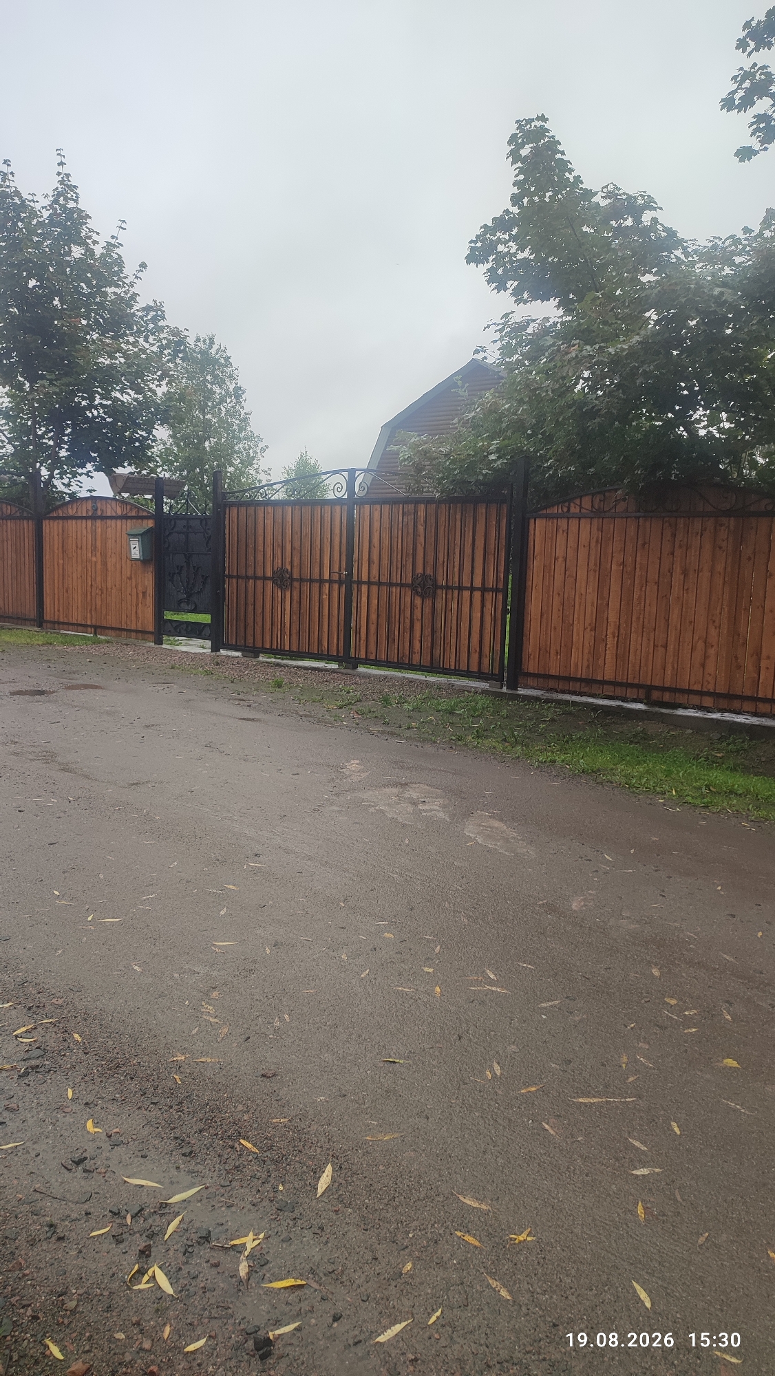 The image depicts a street scene in Suoyarvi, Russia, likely on a cloudy day, given the overcast sky. The foreground is a wet asphalt road with scattered yellow leaves, indicating it might be autumn or that the road has recently been rained on. Beyond the road is a wooden fence with black metal framing and decorative elements. To the left of the fence, a mailbox is visible. Further in the background, a dark metal gate with ornate scrollwork is integrated into the fence. Beyond the fence, trees with green foliage and a portion of a wooden-clad building with a gable roof are visible. The overall impression is of a residential area with a well-maintained entrance. The timestamp "19.08.2026 15:30" is present in the lower right corner of the image. There are no people or animals visible in the frame.