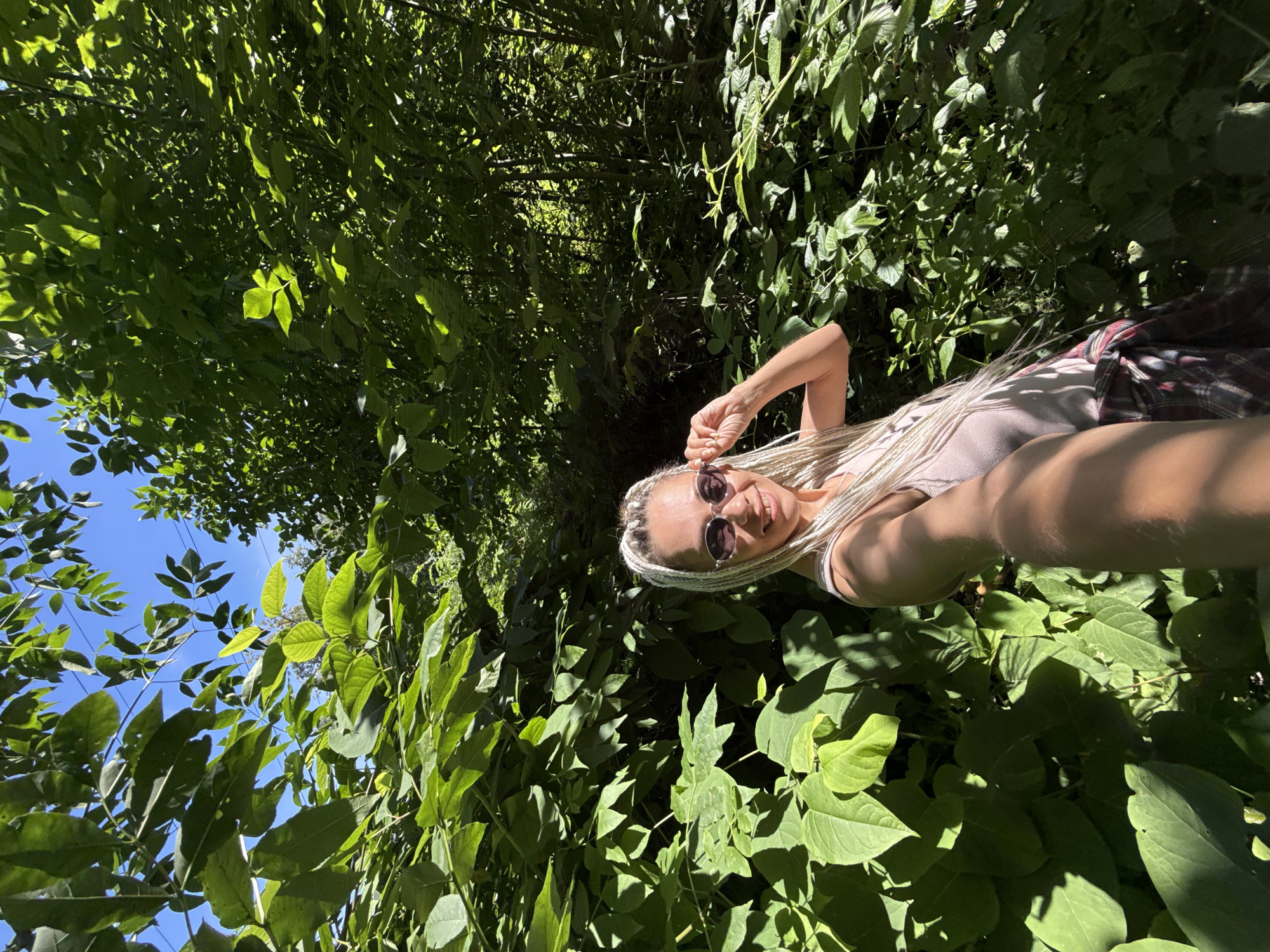 A woman with blonde braided hair and sunglasses is taking a selfie outdoors, surrounded by lush green foliage. She is wearing a pink tank top and a plaid shirt tied around her waist. The setting appears to be a wooded or overgrown area with sunlight filtering through the leaves. The location is identified as Rusanovo, Russia.