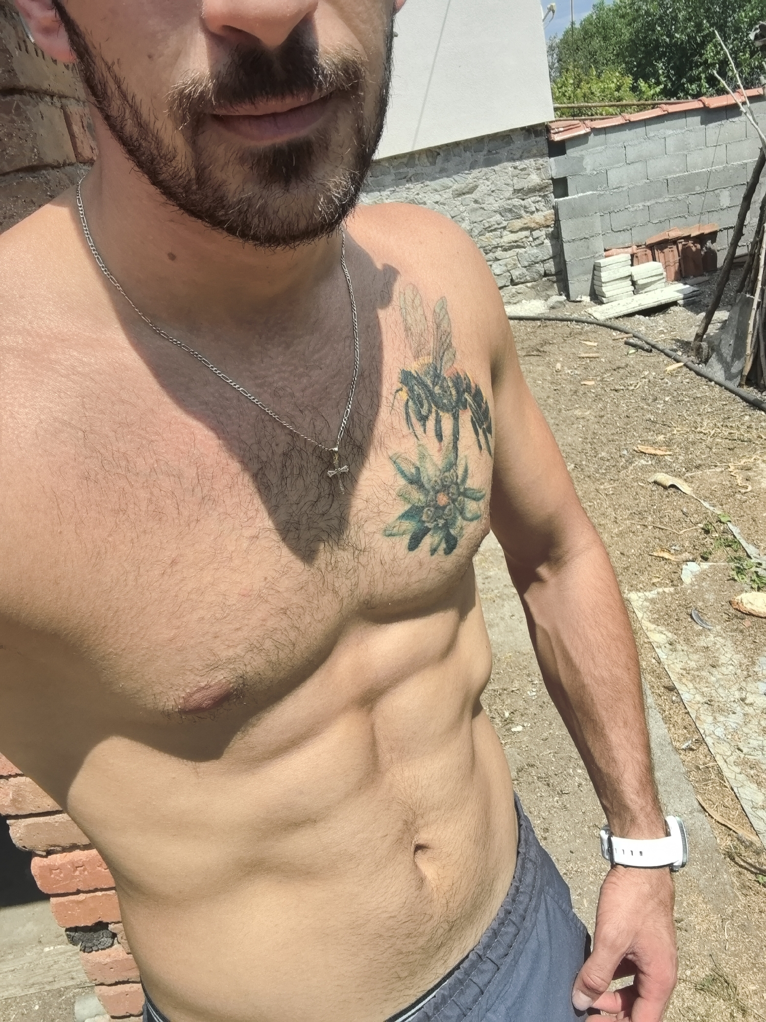 The image shows a man from the chest down, standing outdoors in what appears to be a construction or work area. He has a beard and a tattoo of bees and a flower on his chest. He is wearing a silver chain with a cross pendant and a white wristwatch on his left wrist. His abdomen is well-defined, suggesting he is muscular.
The background shows a partially constructed building with brick and cinder block walls, piles of building materials, and some vegetation. The ground is covered in gravel and dirt. The lighting suggests it is daytime and sunny, with strong shadows cast. The presence of construction materials and unfinished walls indicates this is likely a site under renovation or development, possibly in a rural or semi-rural setting. The mention of Kasnakovo, Bulgaria, places the scene in a specific geographical context within that country.