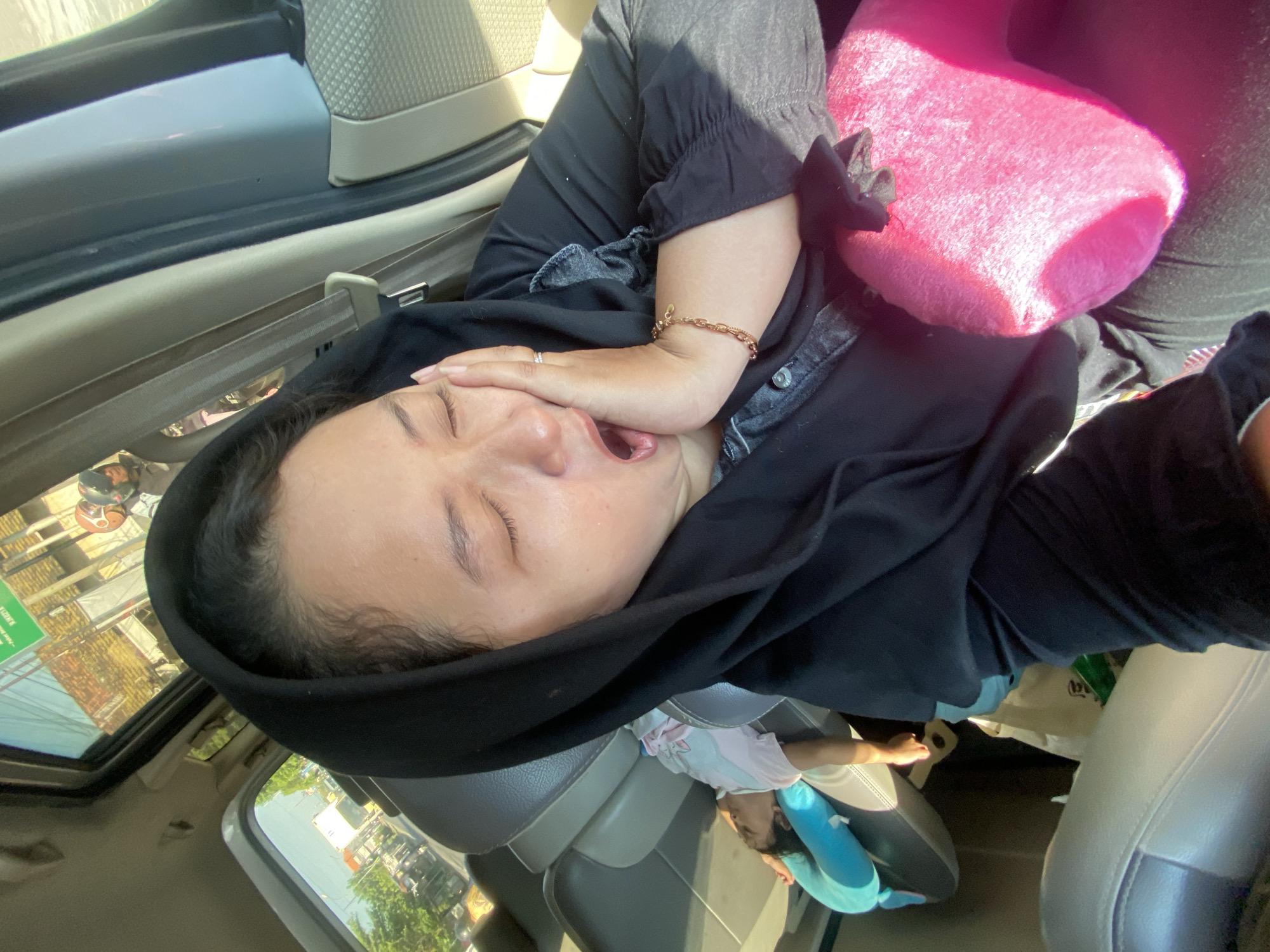 The image depicts a woman in a black hijab yawning inside a vehicle, her hand pressed against her cheek. In the background, a child sleeps on a blue pillow in the car's back seat. The car appears to be in motion, with a view of a street scene through the windows, suggesting a location in the city of Bangsal, Indonesia, possibly on a sunny day. Buildings, trees, and other vehicles are visible outside. A bright pink cushion is held in the woman's lap.