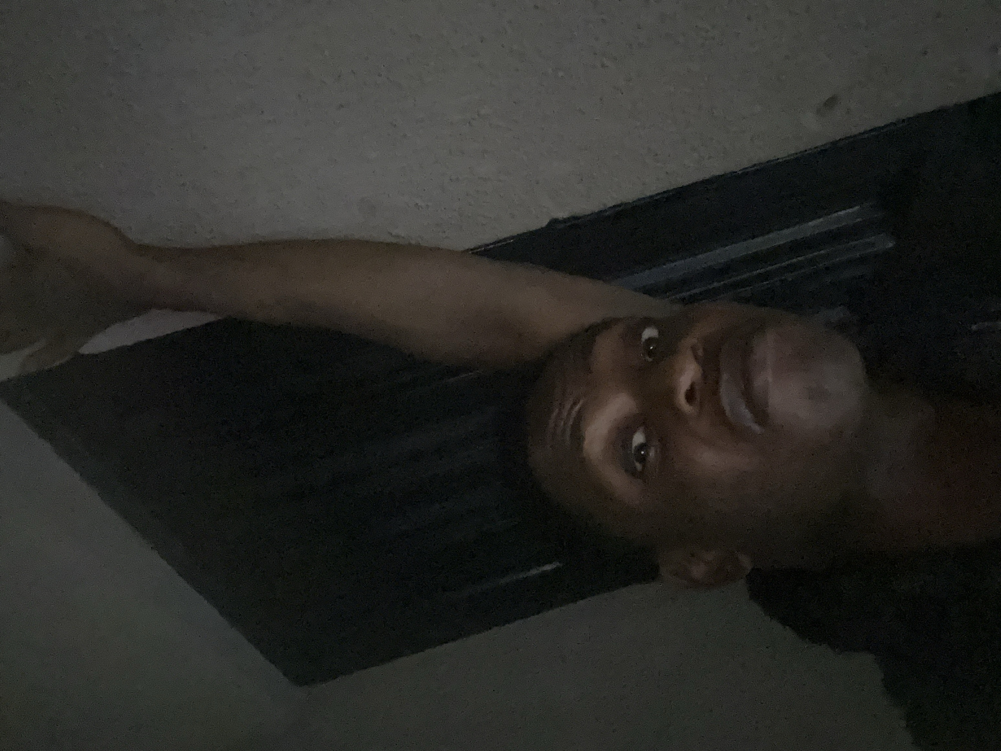 A dimly lit, close-up photograph depicts a man indoors. His face is in the lower-right quadrant of the frame. His left arm extends upwards and leftward, ending with his hand partially visible in the upper-left corner. The background appears to consist of a dark surface and a lighter-colored ceiling. The location is Unknown, Unknown.