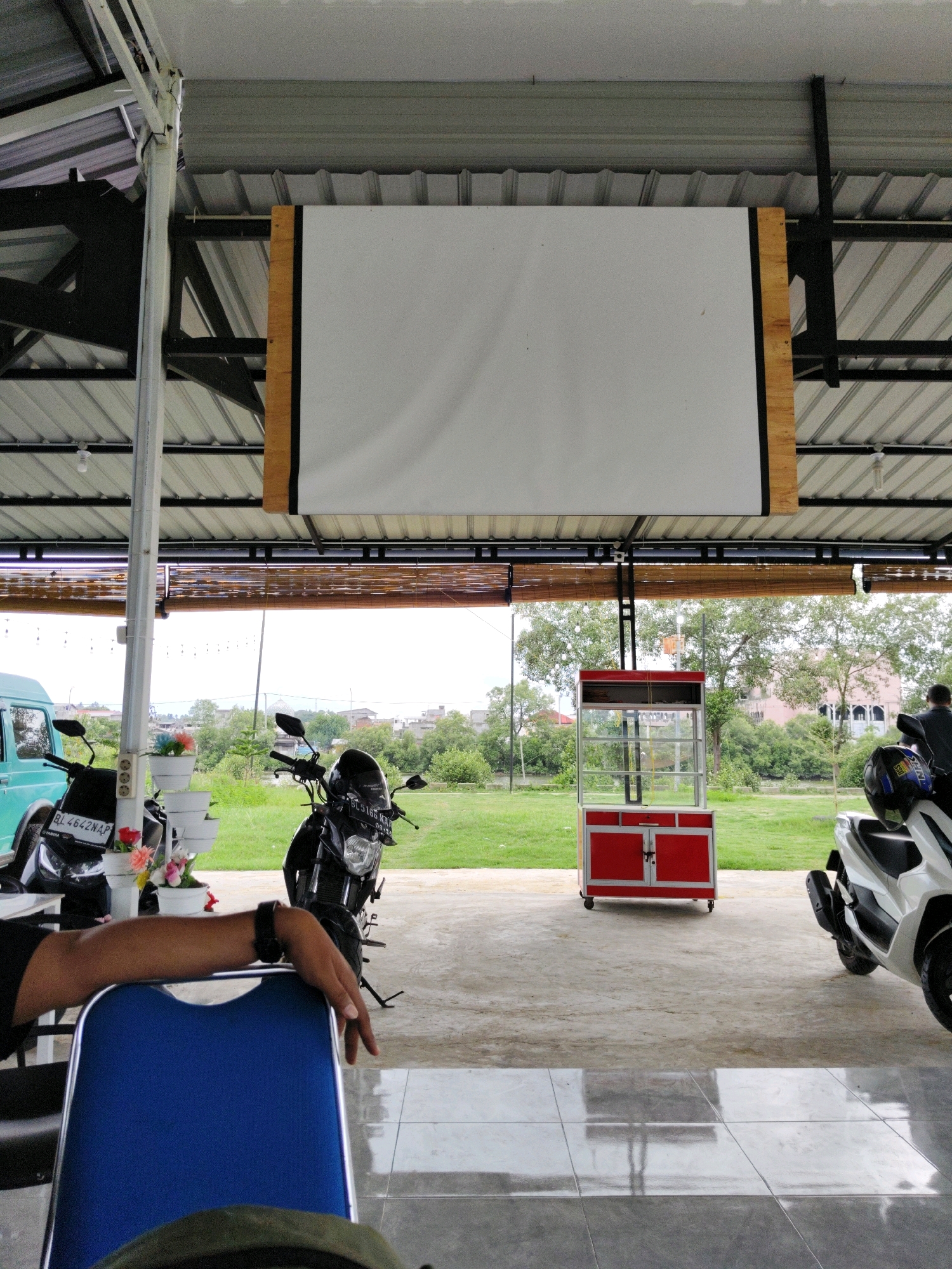 The image captures an outdoor seating area at a cafe or restaurant in Lhokseumawe, Indonesia. The scene is viewed from a seated perspective, looking towards a grassy field with trees in the background. A large, white projection screen is suspended from the roof of the covered area. To the left, a turquoise vehicle is partially visible, and next to it are two motorcycles. One motorcycle, black, has a license plate reading "BL 4642 NAP". A person's arm and hand are resting on a blue chair in the foreground. In the middle ground, a red and silver food cart is visible. A white scooter is parked to the right, with a black helmet placed on its seat. The ground is paved with concrete tiles, which reflect the overhead lighting. The overall atmosphere suggests a casual, open-air establishment.