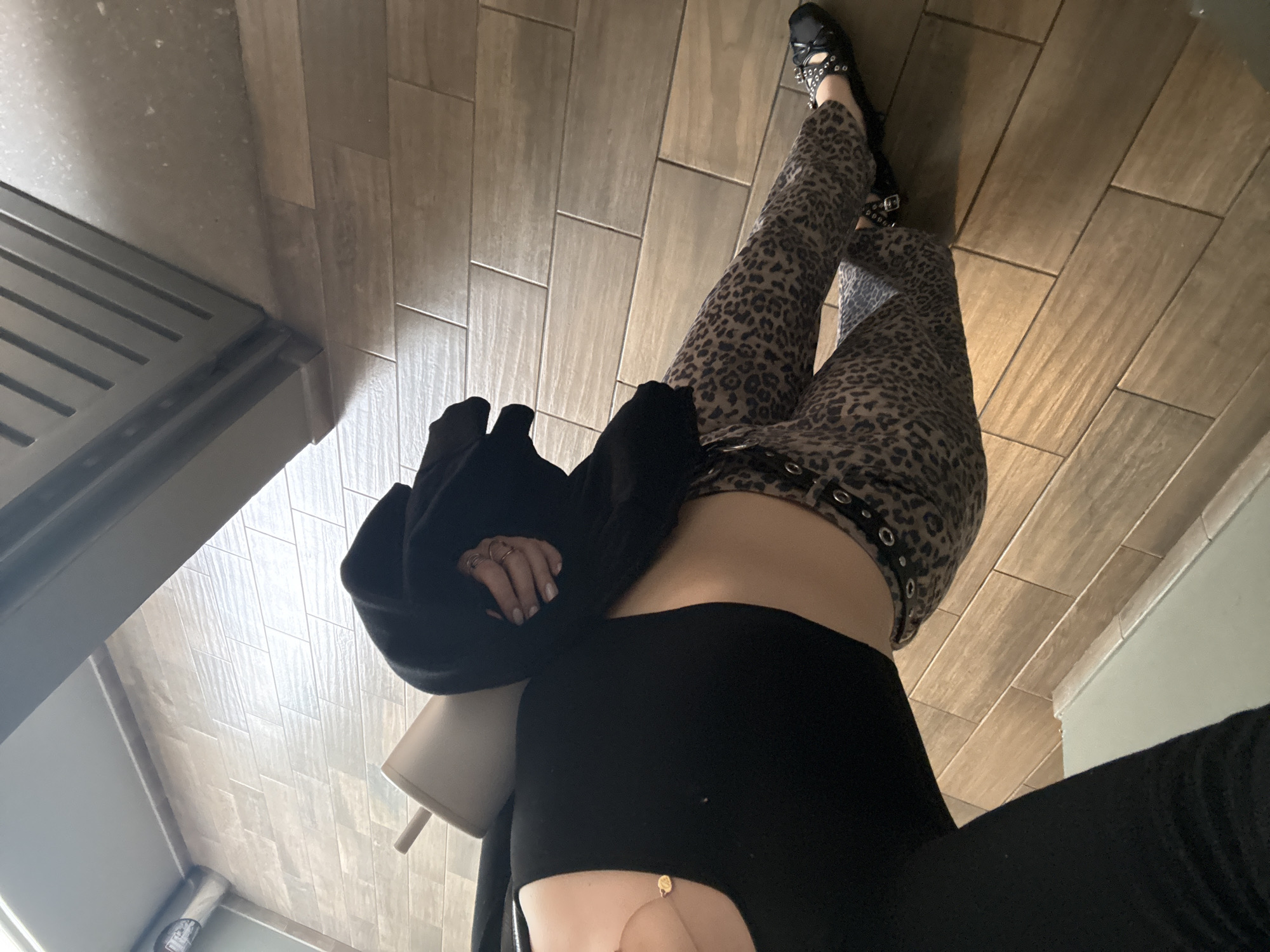 The image, taken from a high-angle perspective, shows a person standing on a tiled floor. The tiles are rectangular and arranged in a herringbone pattern. The individual is wearing leopard-print pants, black shoes with embellishments, and a black top with cut-out details. A black jacket is draped over their arm, and they are holding a white cup with a straw. The setting appears to be indoors, potentially in a commercial space. It is impossible to ascertain whether the location is in Mexico City.