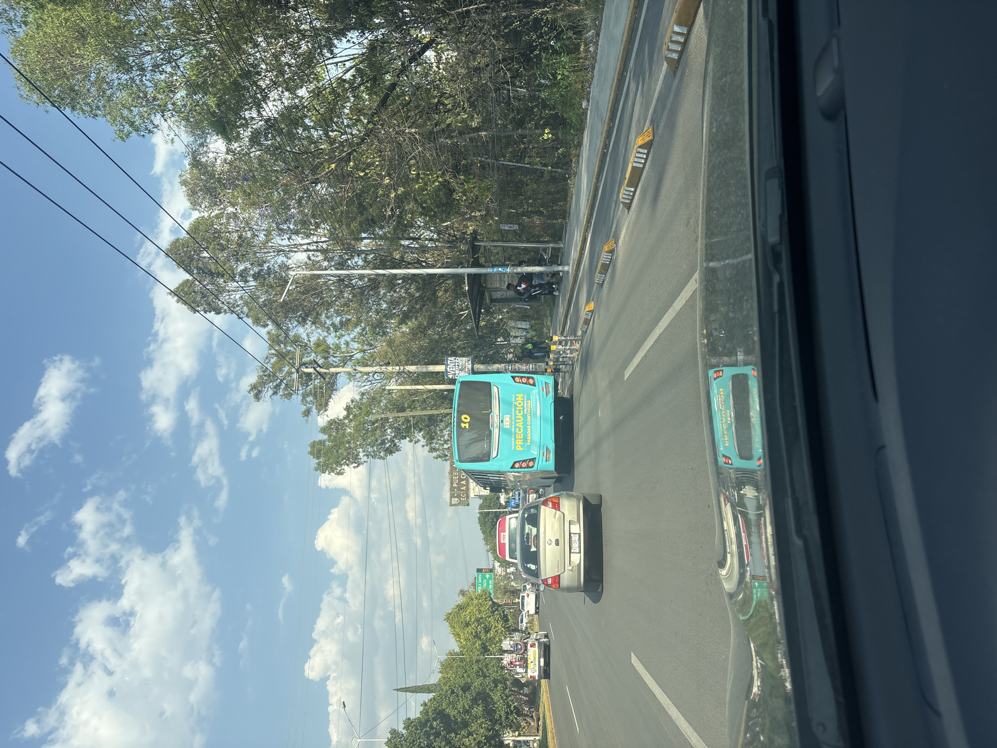 The image captures a street scene in Puebla, Mexico, likely taken from inside a vehicle. The focus is on a multi-lane road with moderate traffic, including a blue public transportation bus with the number "10" prominently displayed. A silver car is directly behind the bus, with other vehicles visible further down the road. Street signs are present, though partially obscured, and trees line the roadside. There is a bus shelter on the sidewalk. Power lines traverse the sky. The weather appears sunny with scattered clouds.