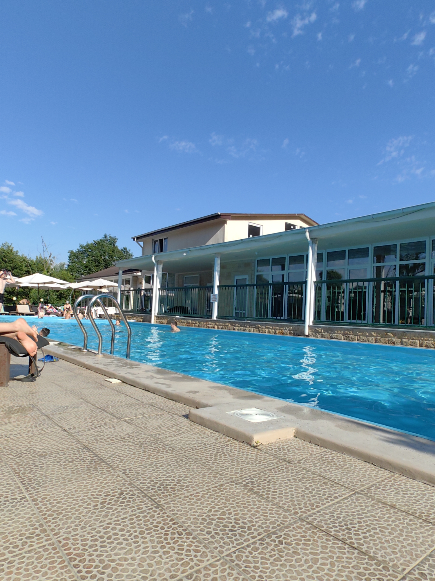 A bright, sunny day at an outdoor swimming pool. The pool is filled with clear blue water and has a set of metal stairs leading into it. The poolside is paved with a tan pebble-like tile. In the background, a two-story building with a light-colored facade and a green railing is visible. There are several people in and around the pool, some swimming, some relaxing on lounge chairs under large white umbrellas. Trees line the edge of the property. The sky is a clear, vibrant blue with a few scattered white clouds. The image appears to be taken in Oryol-Izumrud, Russia.