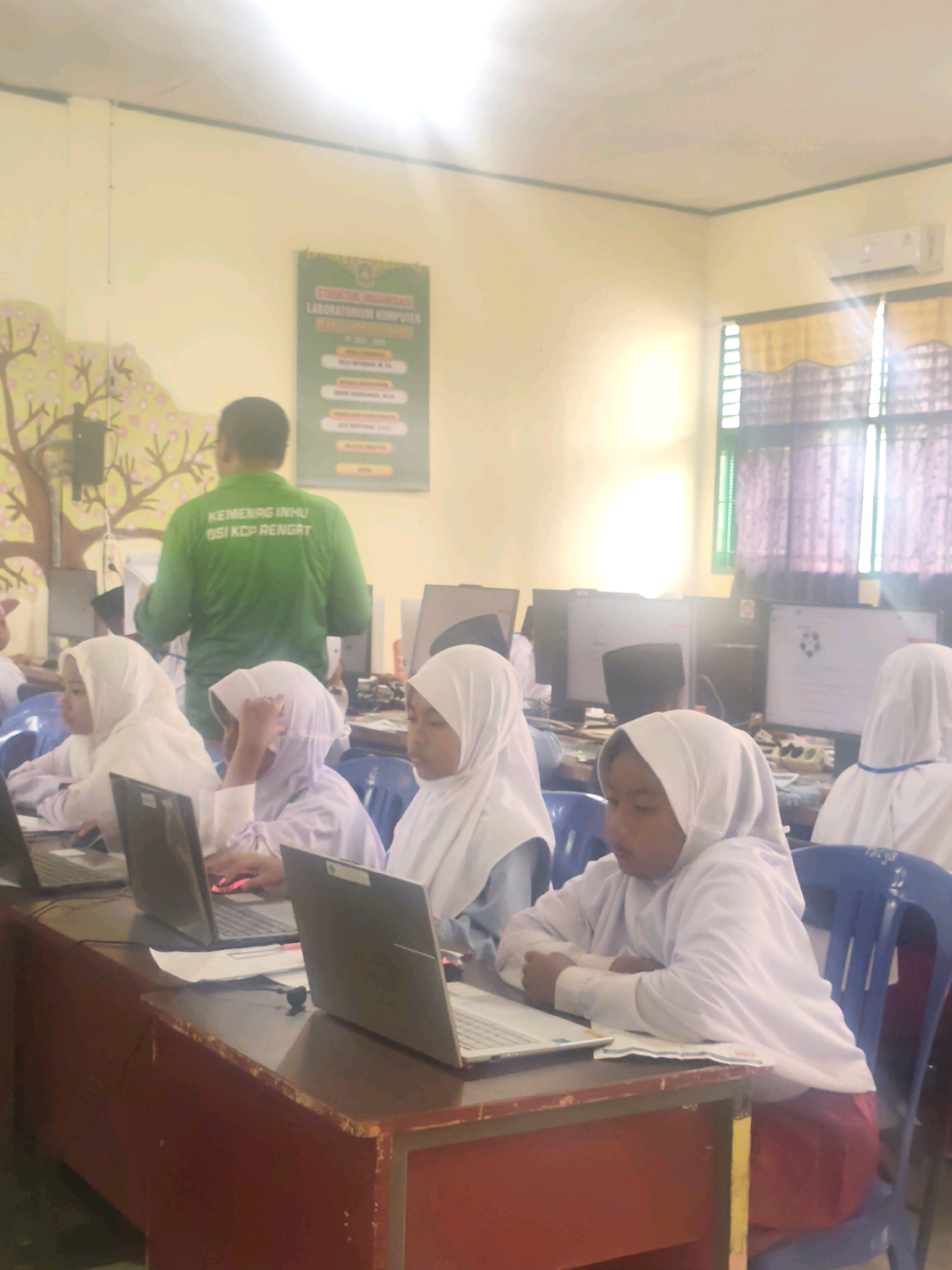 The image depicts a classroom computer lab, likely in Pematang Reba, Indonesia, where several young girls wearing hijabs are engaged with laptops. A man in a green shirt with text reading "KEMENAG INHU" is standing and facing away from the camera, appearing to instruct or supervise. The room features yellow walls adorned with a mural of trees on the left and a poster for a computer laboratory on the right. Multiple computer monitors are visible, some displaying screen content, and the students are seated at desks with open laptops. The environment suggests an educational setting focused on computer literacy, with typical classroom furniture like chairs and desks. The overall lighting suggests it might be daytime, with natural light entering from windows covered by curtains.