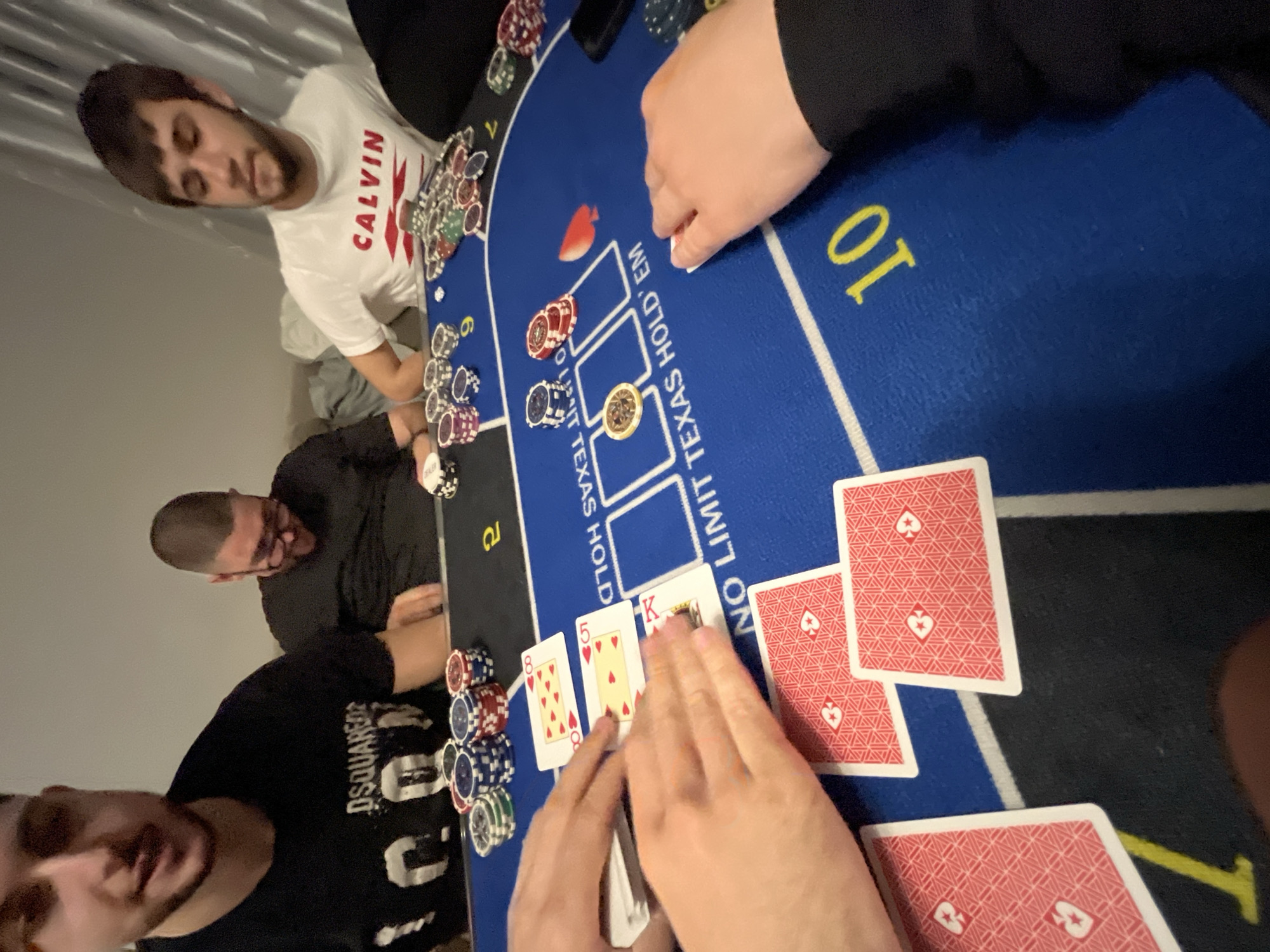 An indoor scene depicts a casual game of Texas Hold'em poker among several men. The primary focus is on a blue poker table with marked player positions and a central area for community cards.
On the table, a "NO LIMIT TEXAS HOLD 'EM" inscription is visible. Poker chips of various colors (red, blue, green, white, and gold) are scattered around, with some stacked in front of players. A white chip marked "DEALER" is present. Three community cards are face-up on the table: the 8 of Hearts, 5 of Hearts, and King of Hearts, indicating the flop has been dealt. Several cards with red backs are also seen, some being held by a player's hands in the foreground, presumably their hole cards or part of the deck.
Several players are visible around the table. One man in a white t-shirt with "CALVIN" printed in red is looking down towards the game. Another man with a buzz cut, wearing a black t-shirt, is also engaged with the game. Partially visible in the foreground is a man wearing a black t-shirt with "DSQUARED2" printed on it. The hands of at least two other individuals are also seen, one seemingly dealing cards.
The lighting suggests it's likely an evening or nighttime game. The atmosphere appears informal and relaxed, typical of a friendly gathering. The game is taking place in Burgas, Bulgaria.
