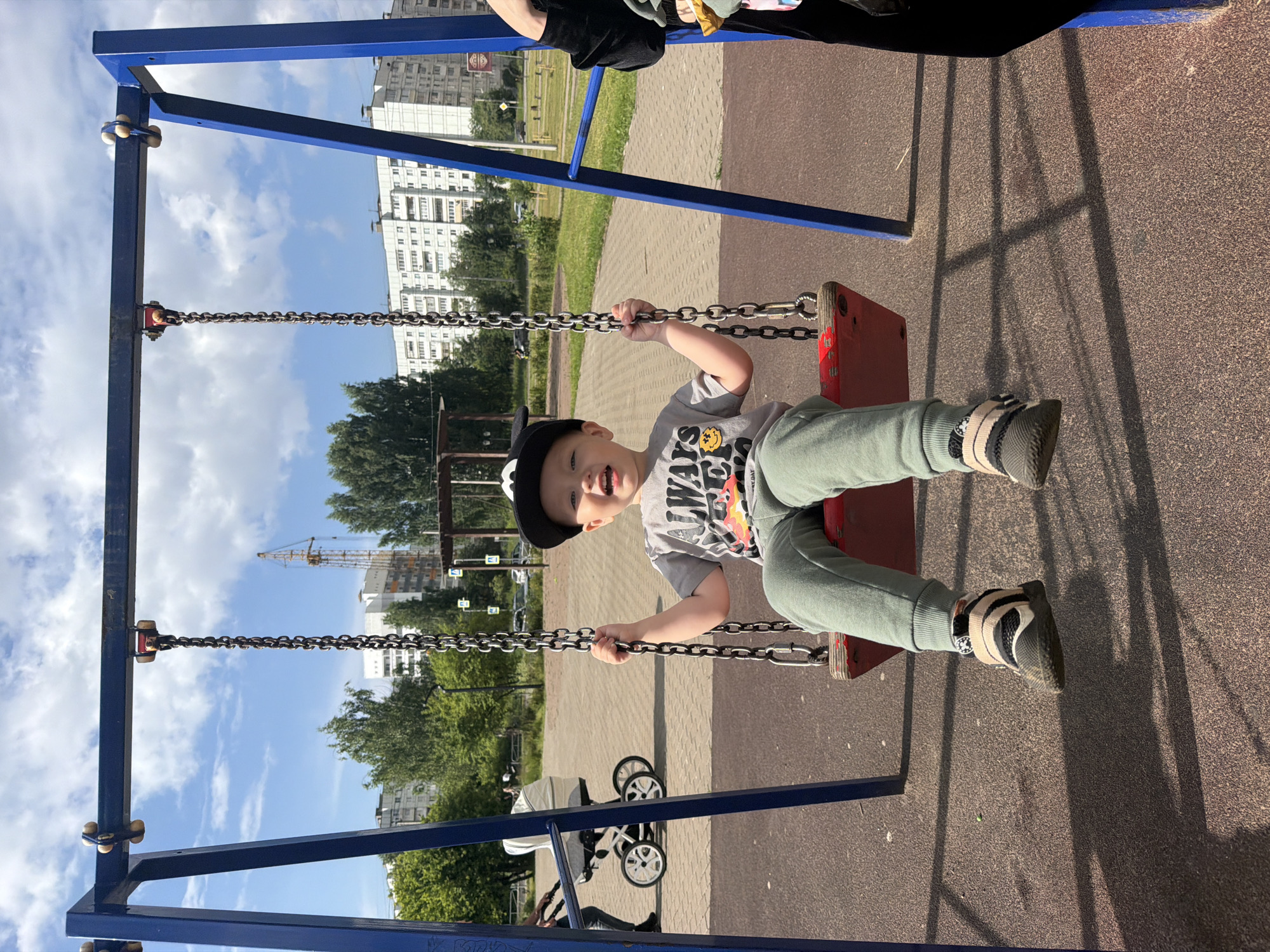 A young boy is sitting on a red swing at a playground in Chaikovsky, Russia. He is wearing a grey t-shirt with "ALWAYS" printed on it, green pants, and black and beige sneakers. He has a black baseball cap on his head. He is smiling and holding onto the swing chains. The playground has a blue metal frame and is located in an urban area with apartment buildings in the background. A stroller and another person are visible to the left of the swing. The ground is covered with reddish-brown material, and shadows from the swing set are cast on the ground. The sky is blue with scattered clouds, and trees are visible in the distance. A crane is also visible in the background.