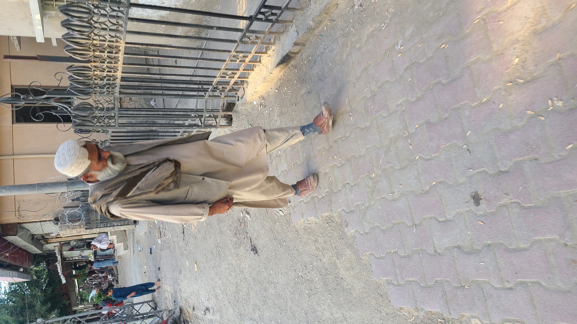 A full-body shot shows an elderly man standing on a sidewalk composed of interlocking pavers. He is wearing a white cap, light brown robe, and brown shoes with visible blue socks. He is facing away from the camera, with his left side partially visible. A dark-colored wrought-iron fence stands between the man and a building. In the background is a street scene with other individuals, including at least one woman in a blue outfit. The location is identified as Quetta City Tehsil, Pakistan.