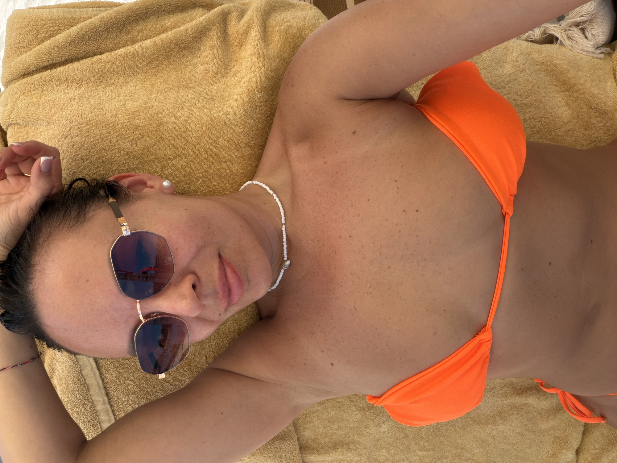 A woman lies on a tan towel, presumably on a beach. She wears an orange bikini and sunglasses. A white beaded necklace adorns her neck, and a small red bracelet encircles her left wrist. An earring is visible in her left ear. The background includes a partially visible white fabric with knotted fringe, lying on the towel. The location is Bac Nha Trang Ward, Vietnam.