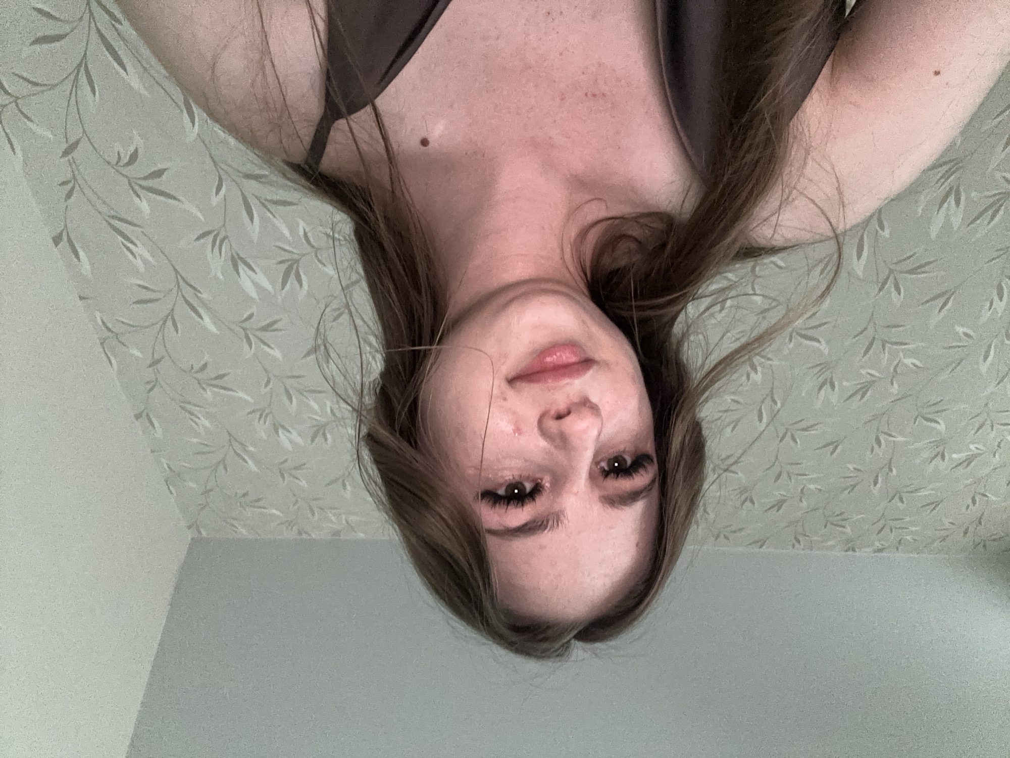 The image shows a close-up, low-angle shot of a young woman's face and upper body. She has long, brown hair that is slightly windswept and appears to be wearing a dark-colored, silky garment with thin straps. Her eyes are looking slightly to her left, and her lips are slightly parted. She has on false eyelashes and her skin has some freckles and blemishes. The background consists of a wall with light-colored wallpaper featuring a repeating pattern of thin, branch-like motifs. The ceiling is visible at the top of the frame. The lighting is soft, possibly from an overhead source. The image is taken indoors, likely in a residential setting, with no indication of an urban environment or a specific city.