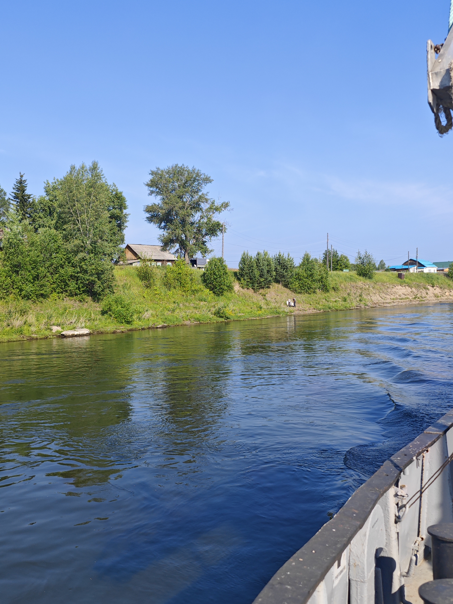 The image shows a wide river with a boat in the foreground, viewed from its starboard side. The river water is dark blue with rippling waves. On the opposite bank, a grassy slope rises to a treeline with several houses and structures. The sky is clear and blue. In the middle ground of the far bank, a small group of people, appearing to be two adults and one child, are standing near the water's edge. The scene depicts a rural or suburban setting along a river, potentially in or near the village of Galanino, Russia, given the context. The time of day appears to be daytime, likely late morning or afternoon, due to the bright sunlight.