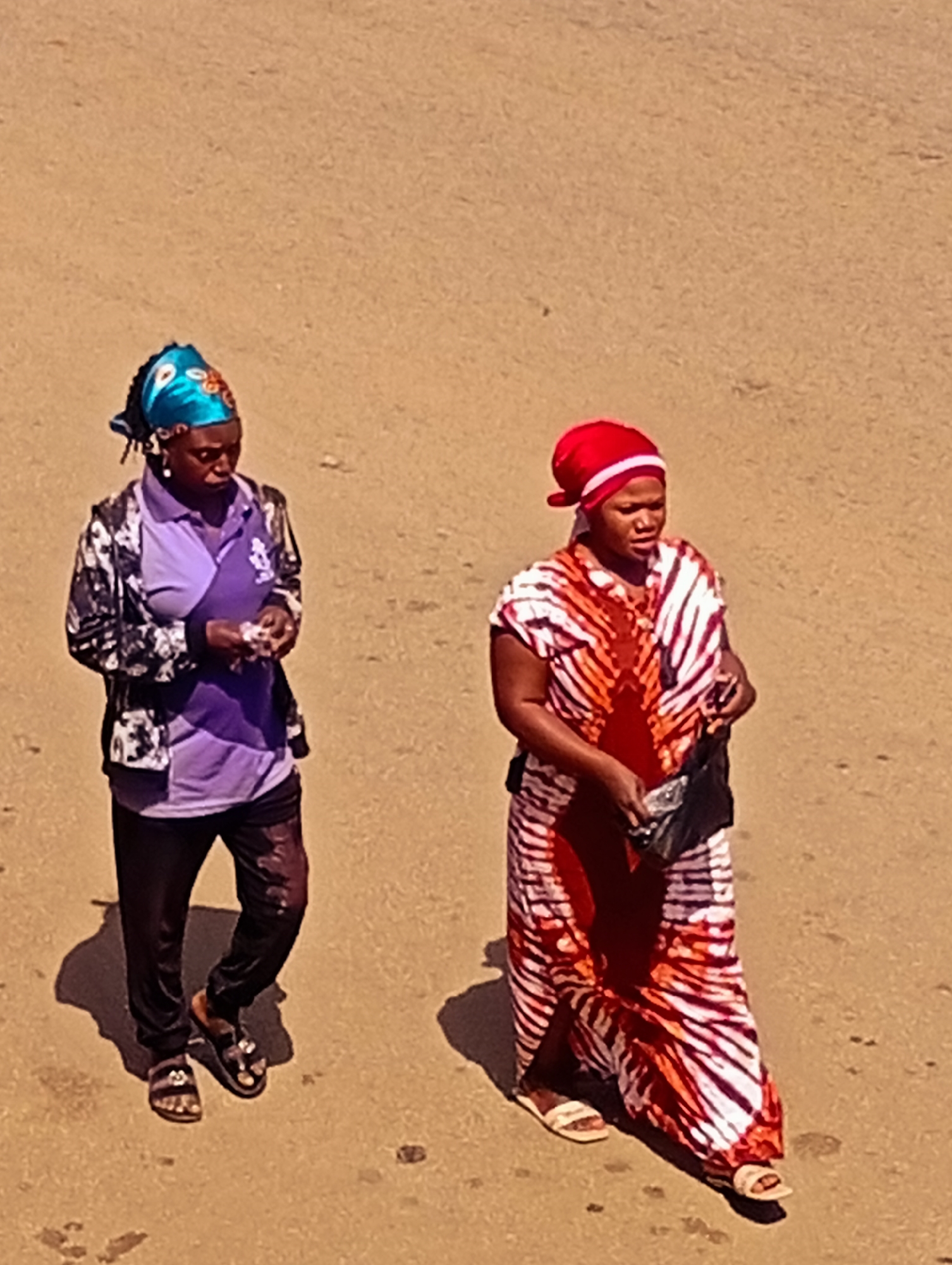 In Bukuru, Nigeria, two women are walking on a sandy surface. The woman on the left wears a vibrant turquoise headscarf with a floral pattern and a purple polo shirt under a black and white patterned jacket. She has black pants and is wearing black sandals. She is looking down and holding small items in her hands. The woman on the right has a red headscarf with a white stripe and a colorful dress with red and white tie-dye patterns. She is carrying a black bag in her right hand and is wearing light-colored sandals. Both women are walking from left to right. The ground is a light brown, sandy terrain with scattered small dark specks. The lighting suggests it is daytime, casting distinct shadows to the left of the women.