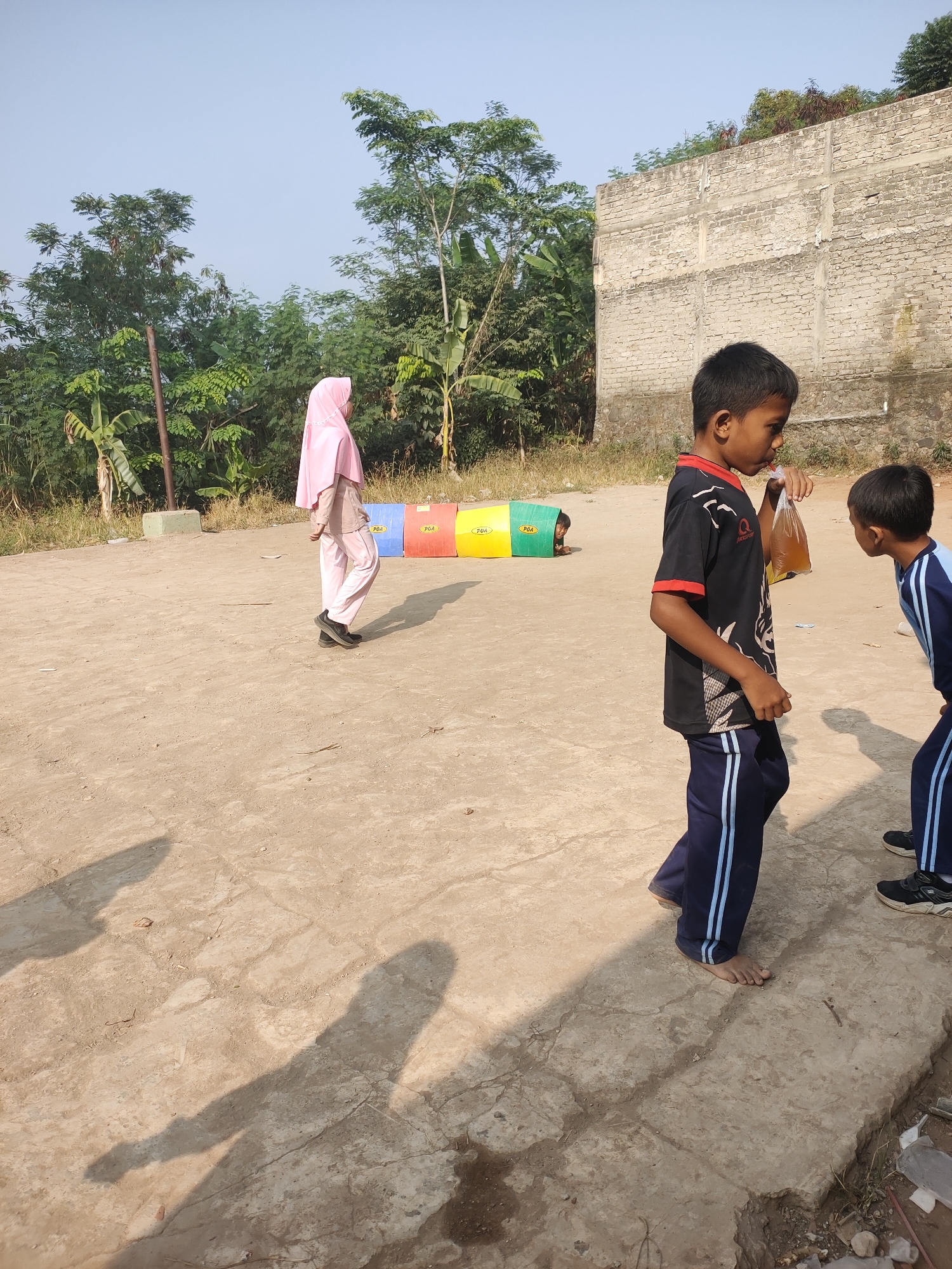 In Subang, Indonesia, on a bright, sunny day, two young boys are the central figures on a dirt and concrete playground. The boy on the left, wearing a black t-shirt with red trim and dark blue pants with white stripes, is drinking from a clear plastic bag filled with a light brown liquid. He is facing right and appears to be looking at the other boy. To his right, another boy in a blue and white striped uniform is partially visible, also facing the first boy. In the background, a young girl wearing a light pink hijab and matching clothing walks away from the camera. Behind her are four brightly colored cylindrical objects, red, yellow, blue, and green, possibly part of a play structure or obstacle course. Further in the background, lush green trees and a weathered concrete wall are visible, suggesting an outdoor area adjacent to a building. The ground is a mix of packed dirt and cracked concrete, with shadows cast by the subjects and the surrounding environment.