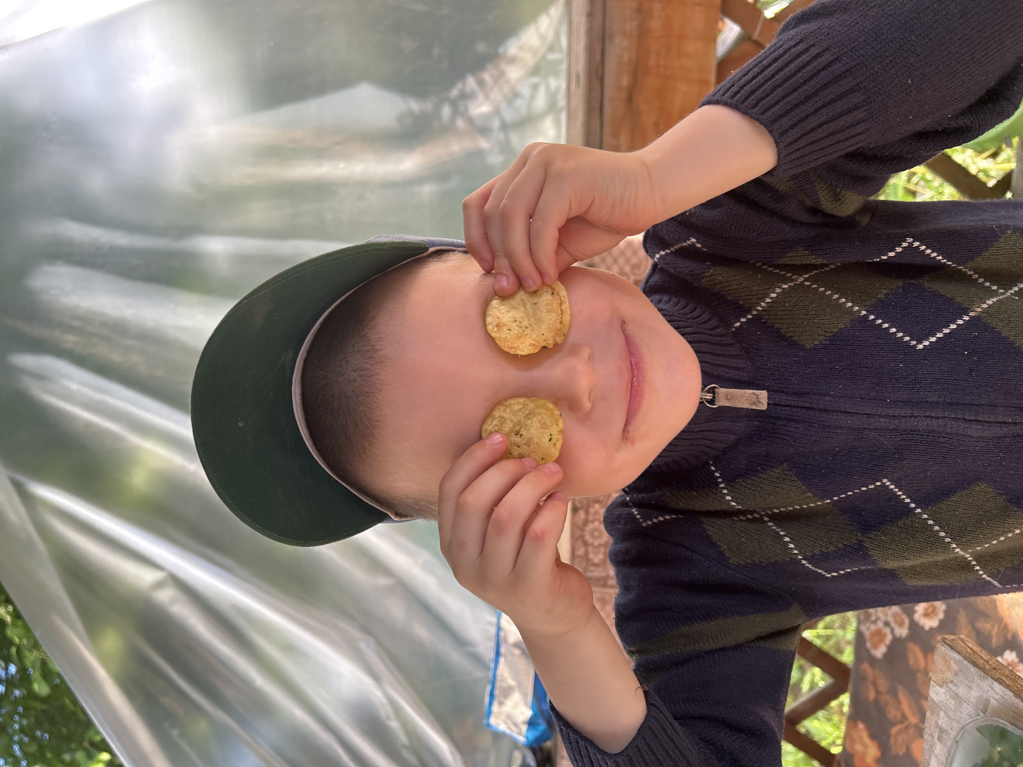 A young male child, approximately 5-8 years old, is centrally positioned, looking forward with a slight smile. He is holding a single round potato chip over each eye with his hands, obscuring his vision. The child wears a dark green baseball cap, reversed with the brim facing backward, and a navy blue zip-up sweater featuring an argyle pattern of dark green and white diamonds. The zipper is visible on the front of the sweater. The scene takes place outdoors in the city of Фоки, Russia. The background consists of a large, translucent, wrinkled sheet of material, likely plastic or polyethylene, suggesting a temporary covering such as a tent or gazebo. Through this material, soft green shapes indicative of foliage and hints of bright sky are visible. The child is seated at a light-colored wooden table, parts of which are visible in the lower left and right foreground. A partially obscured blue and white bag is visible on the surface to the left of the child's body. The illumination is natural daylight.